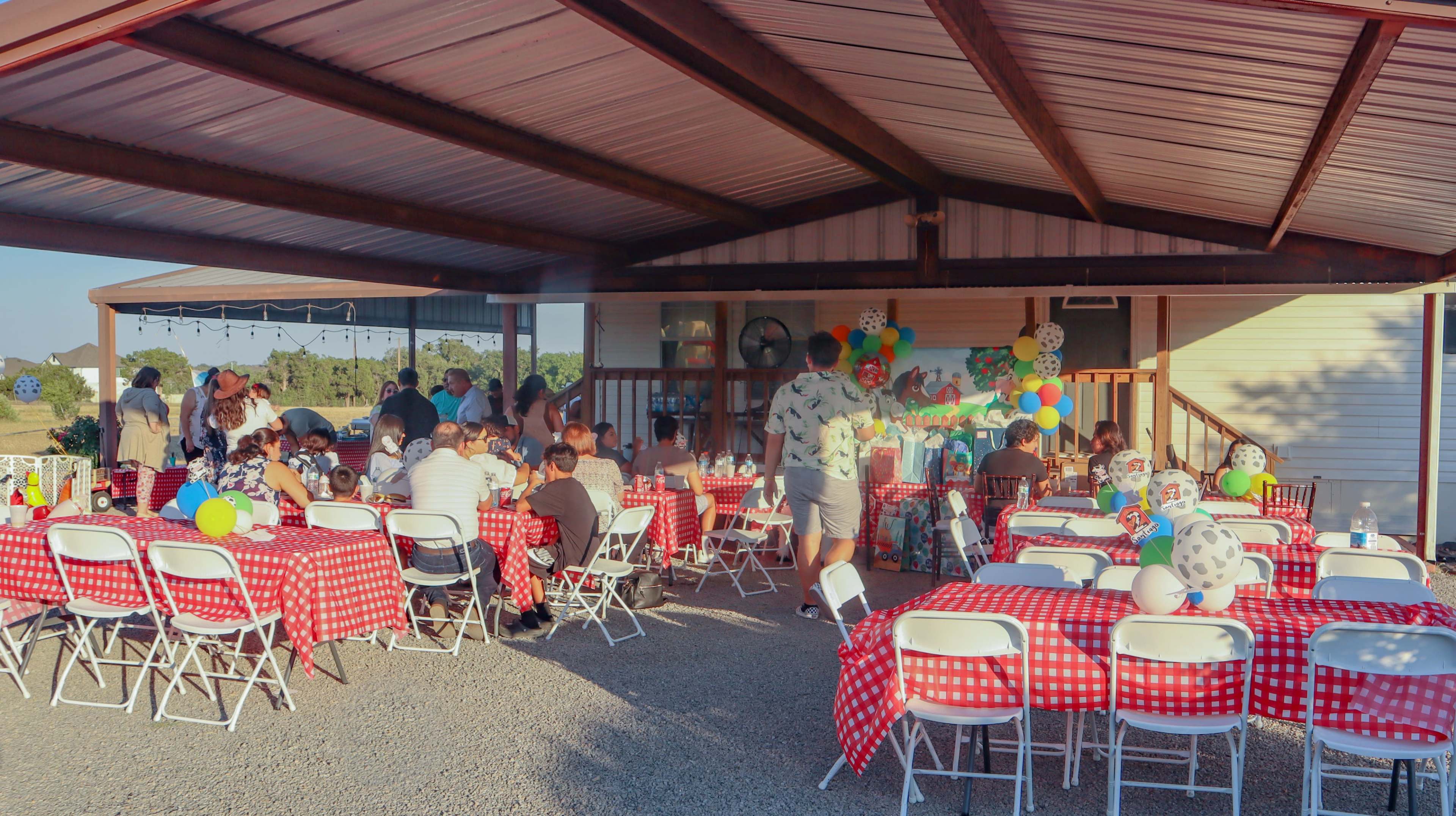 A large outdoor gathering is set up under a covered area, featuring tables with red-checkered tablecloths, people mingling, and colorful decorations.