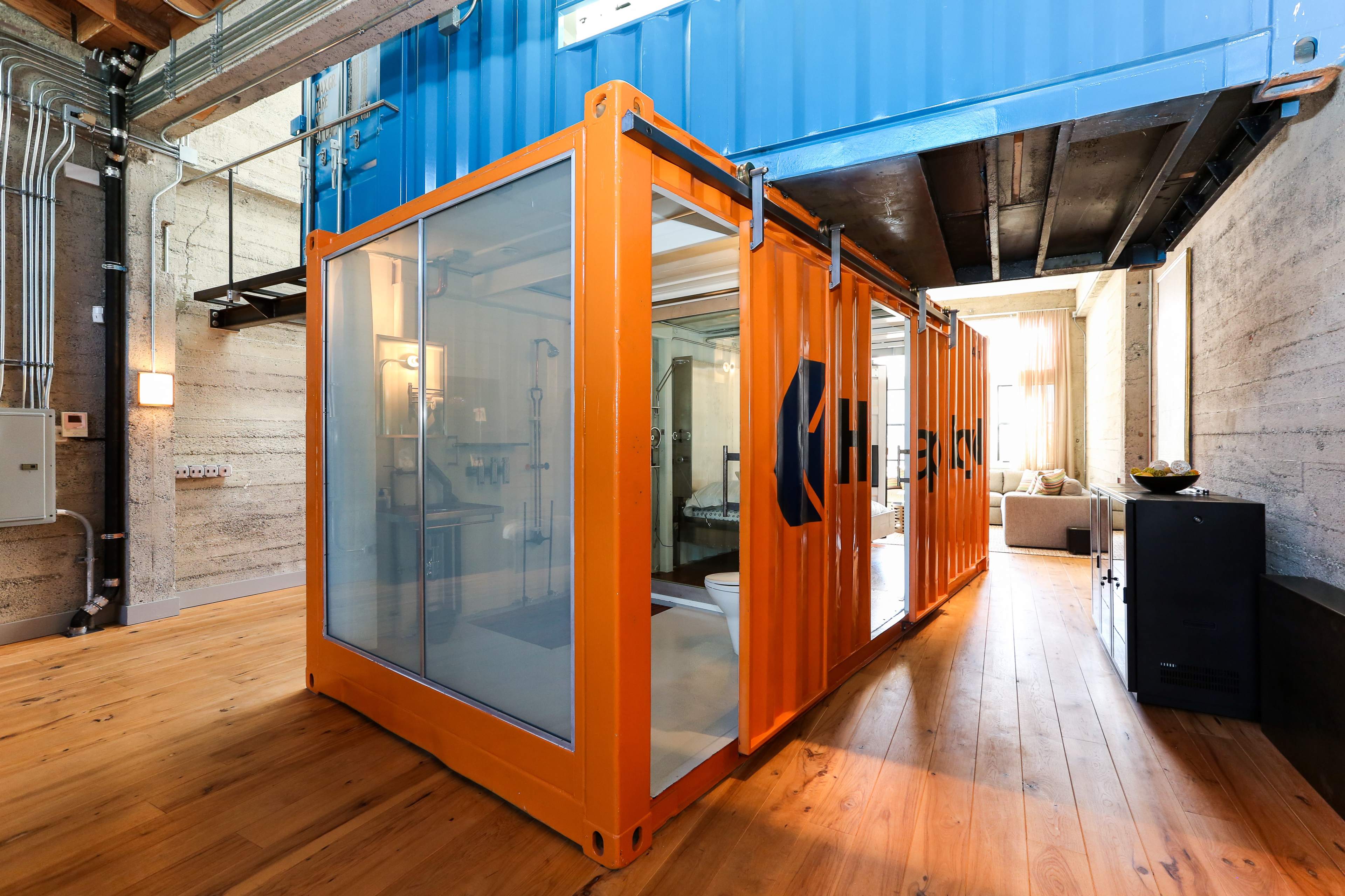 A bright, modern space features an orange shipping container converted into a bathroom, surrounded by wood flooring and industrial-style architecture.