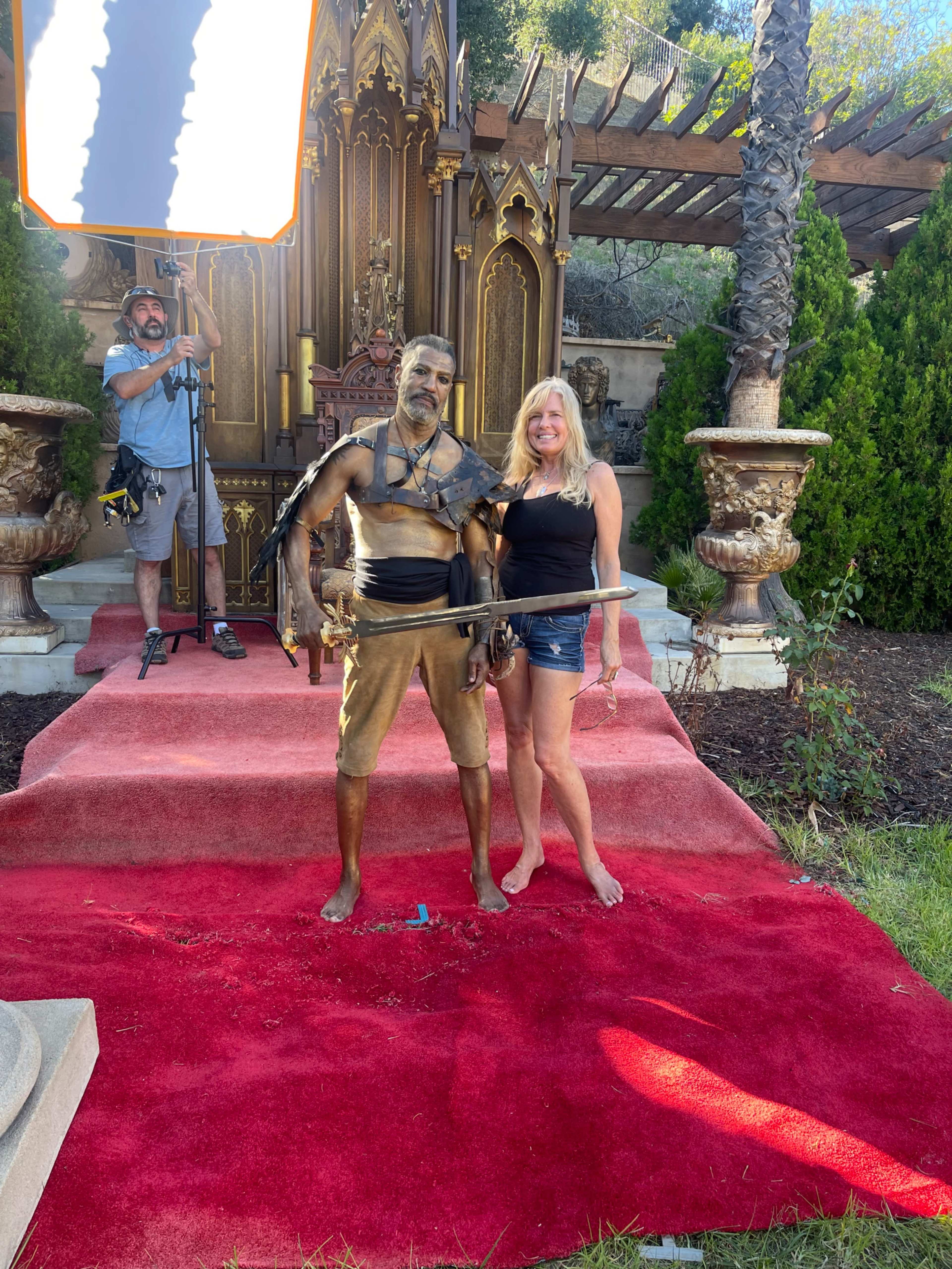 A woman stands barefoot on a red carpet next to a man dressed in bronze armor with a sword, while a crew member operates a light in the background.