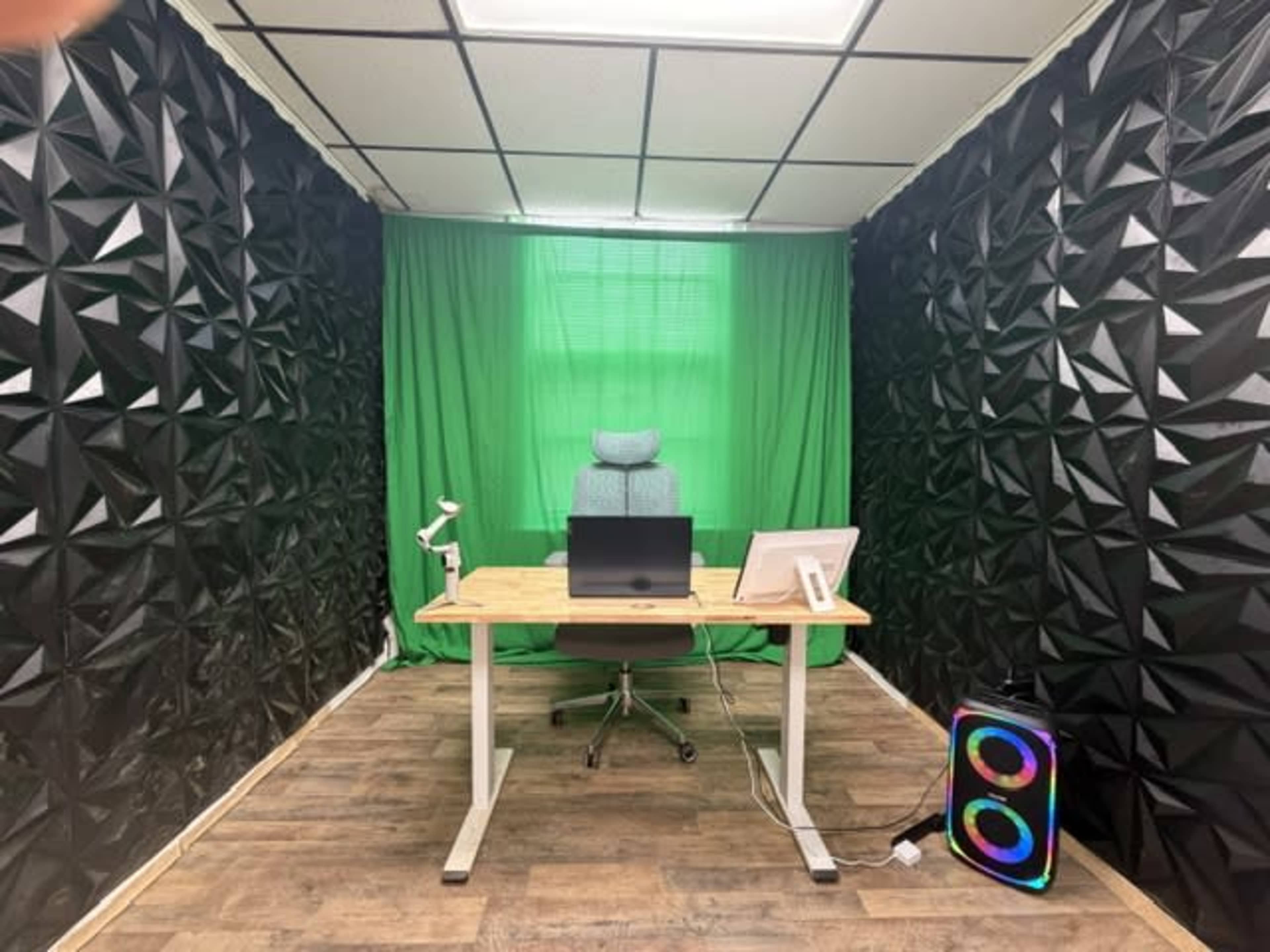 The image depicts a home office with a desk, computer setup, and a green backdrop, surrounded by textured black walls.