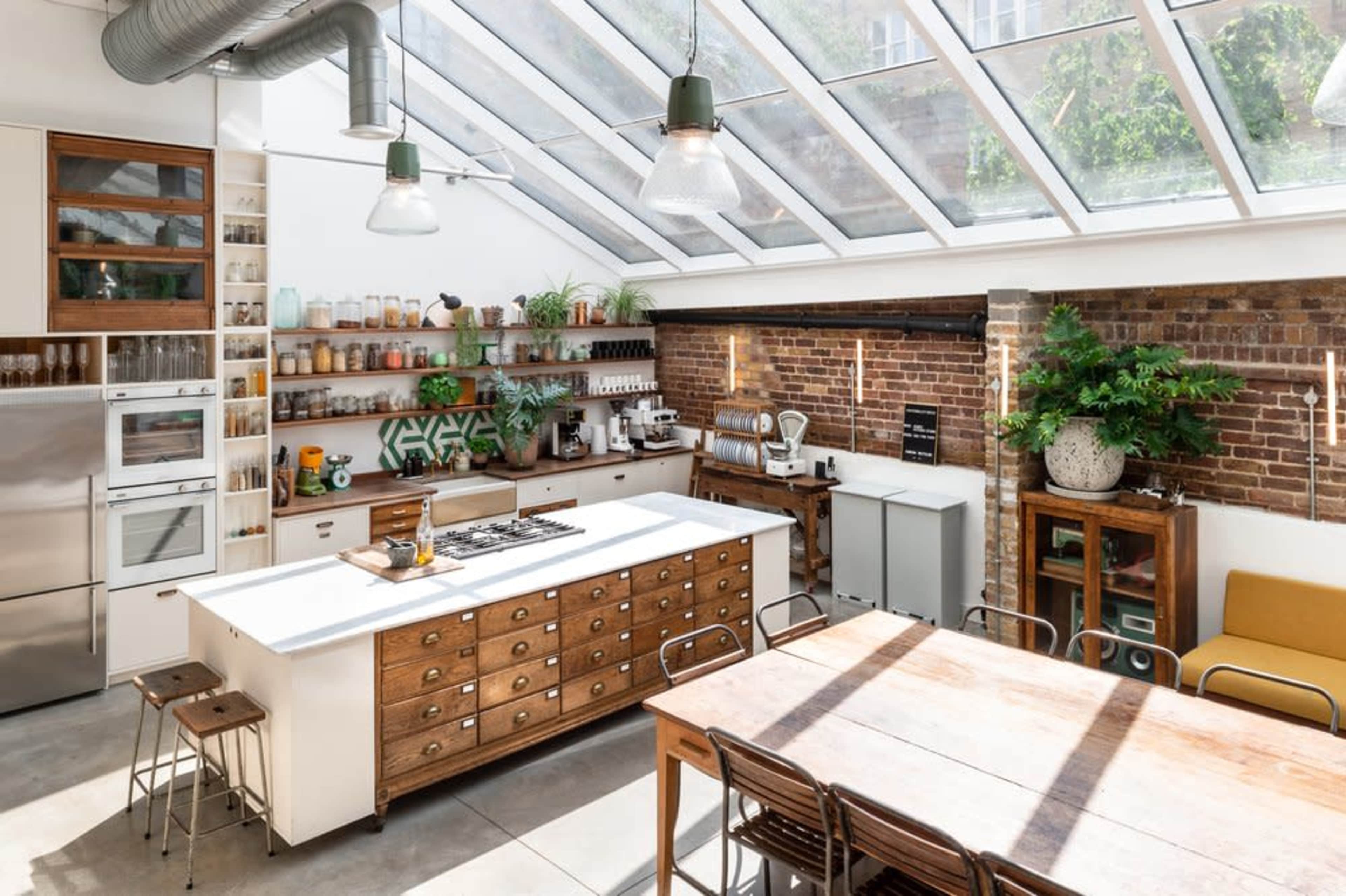A modern kitchen features a large central island with wooden drawers, a wall of shelves filled with jars, and a glass roof that illuminates the space.