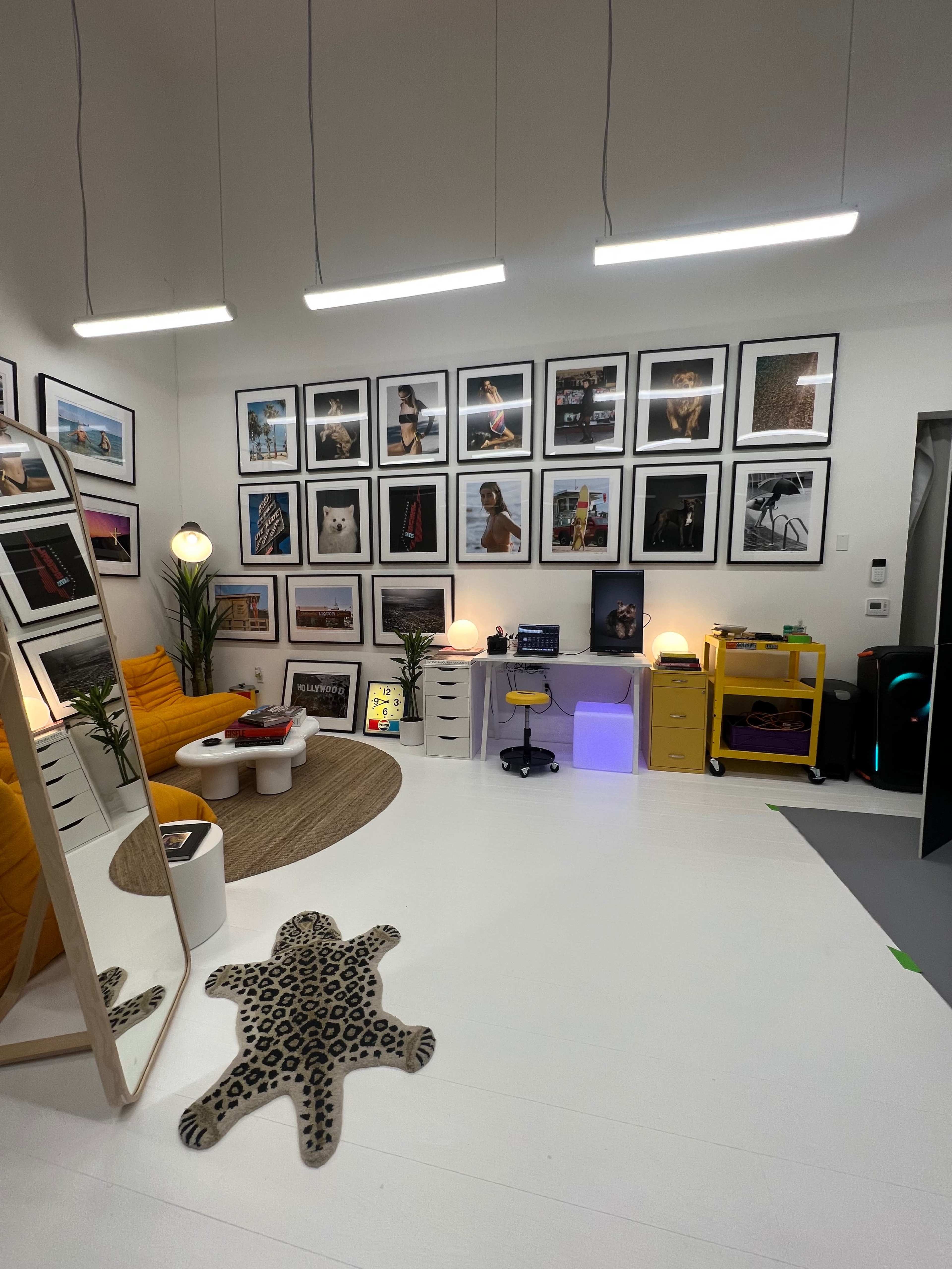 A stylish room featuring a yellow couch, a large mirror, various framed photographs on the wall, and a mix of furniture including a desk and storage units.