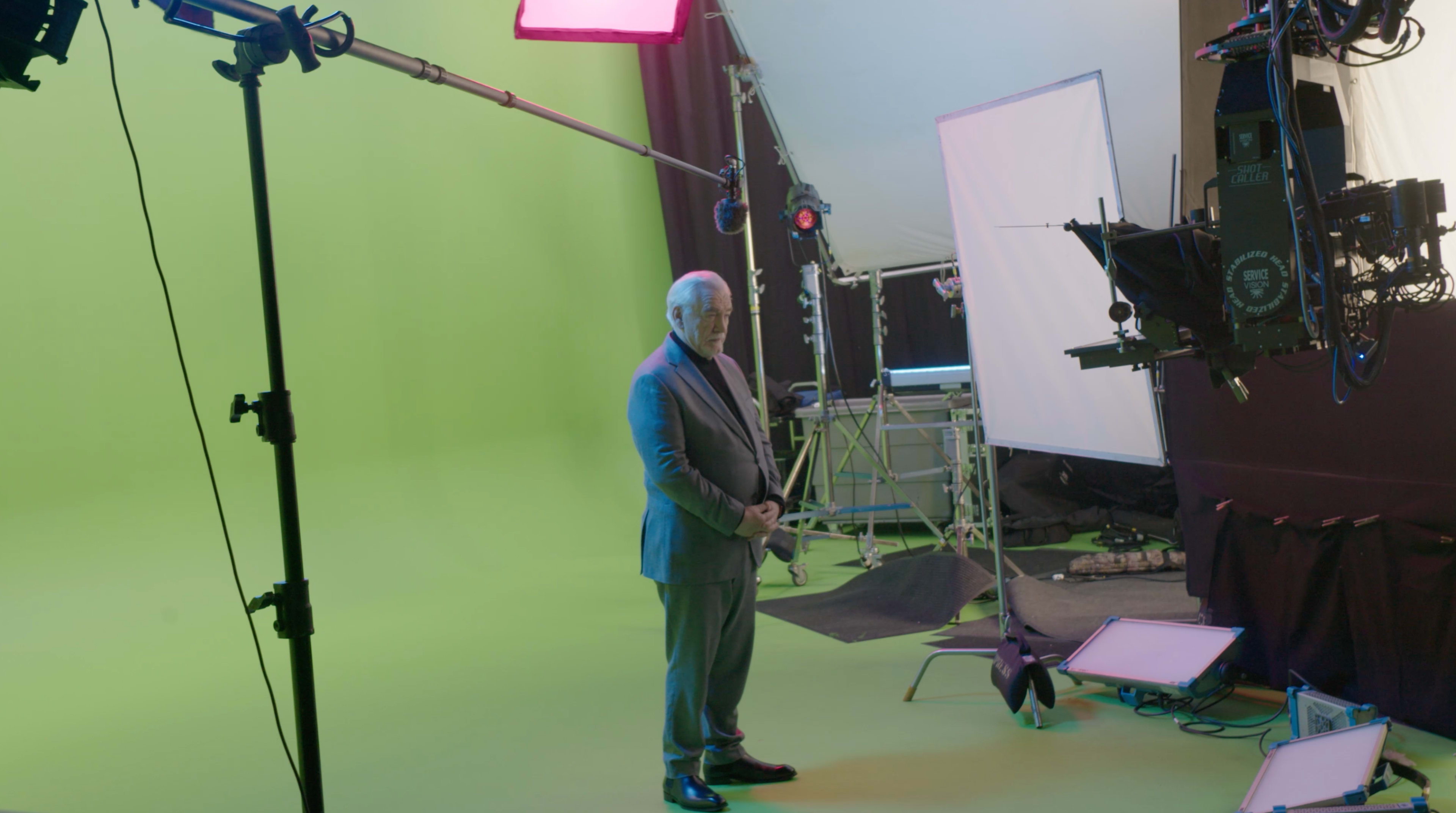 A man in a suit stands in a green screen studio surrounded by lighting equipment and cameras.