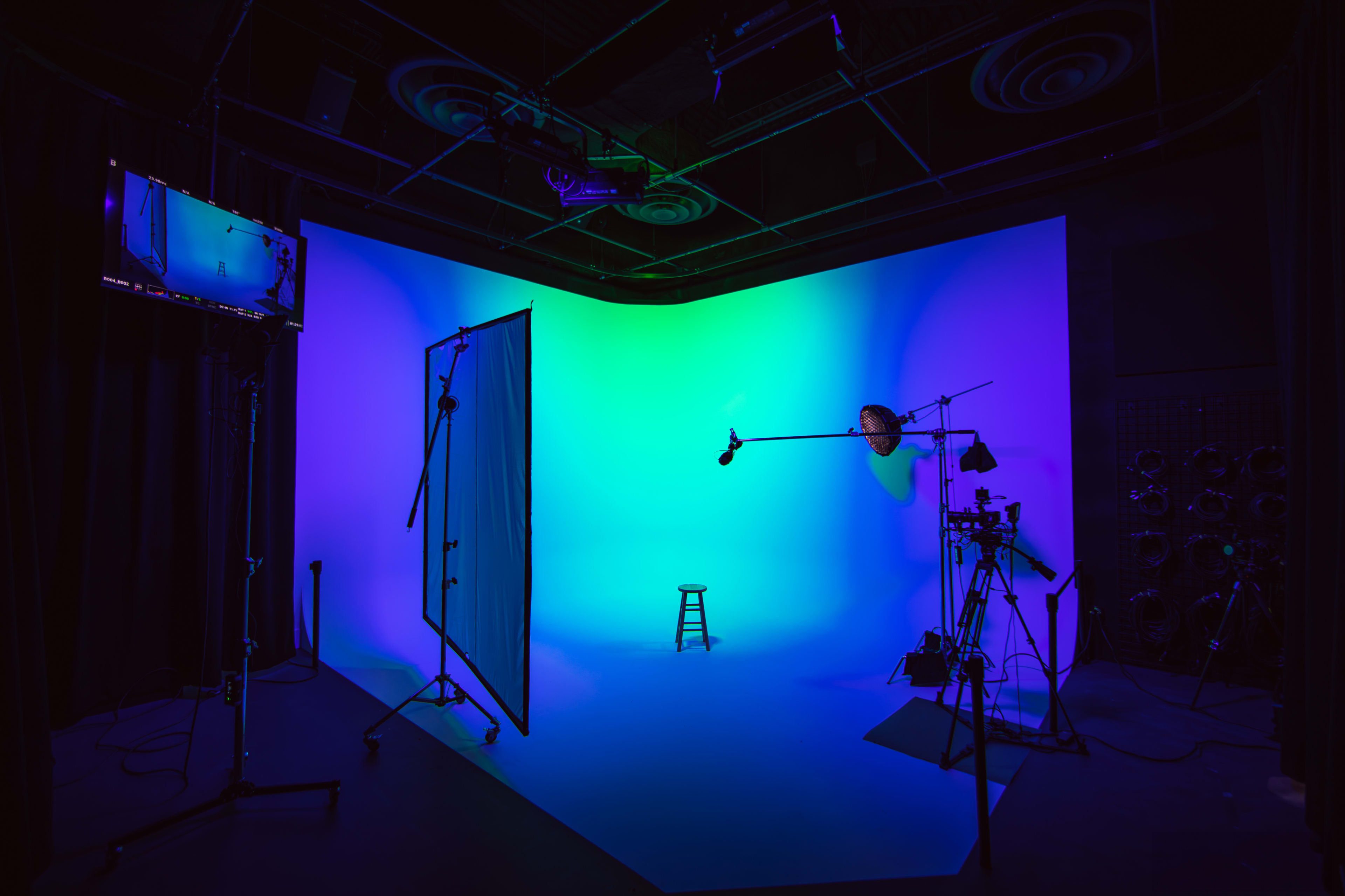 A photography studio with a blue and green backdrop, a stool in the center, and various lighting and camera equipment set up around the area.