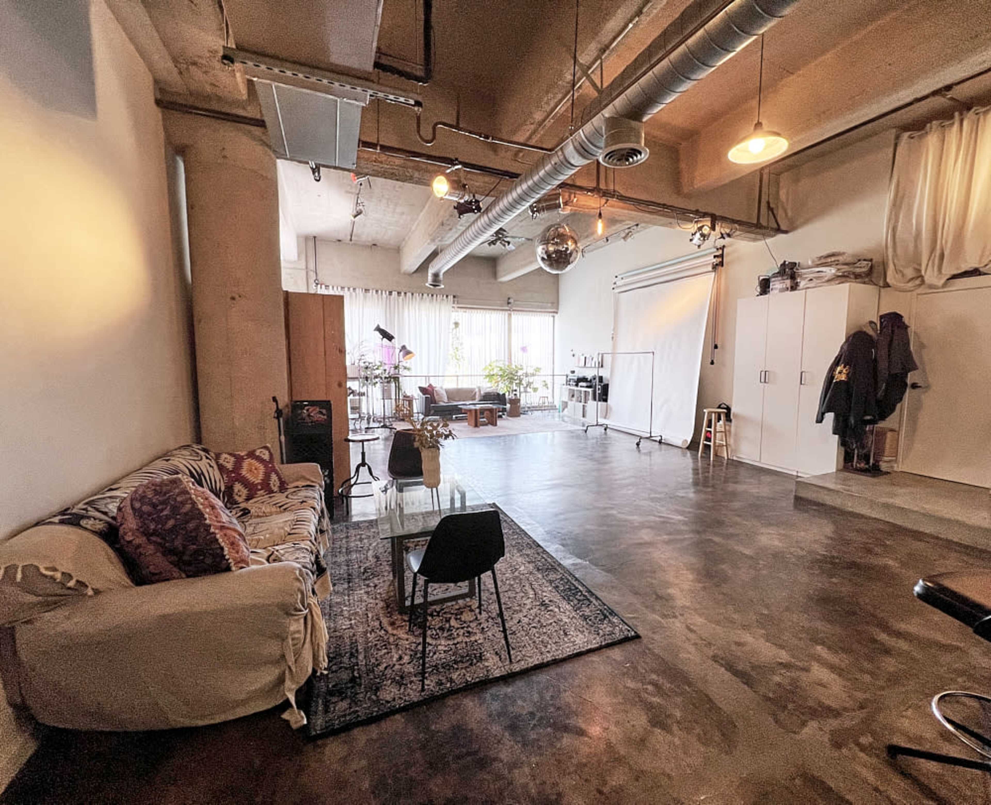 A spacious studio with a concrete floor, a large window, a couch, a chair on a rug, and various furnishings including a mirror ball and storage cabinets.