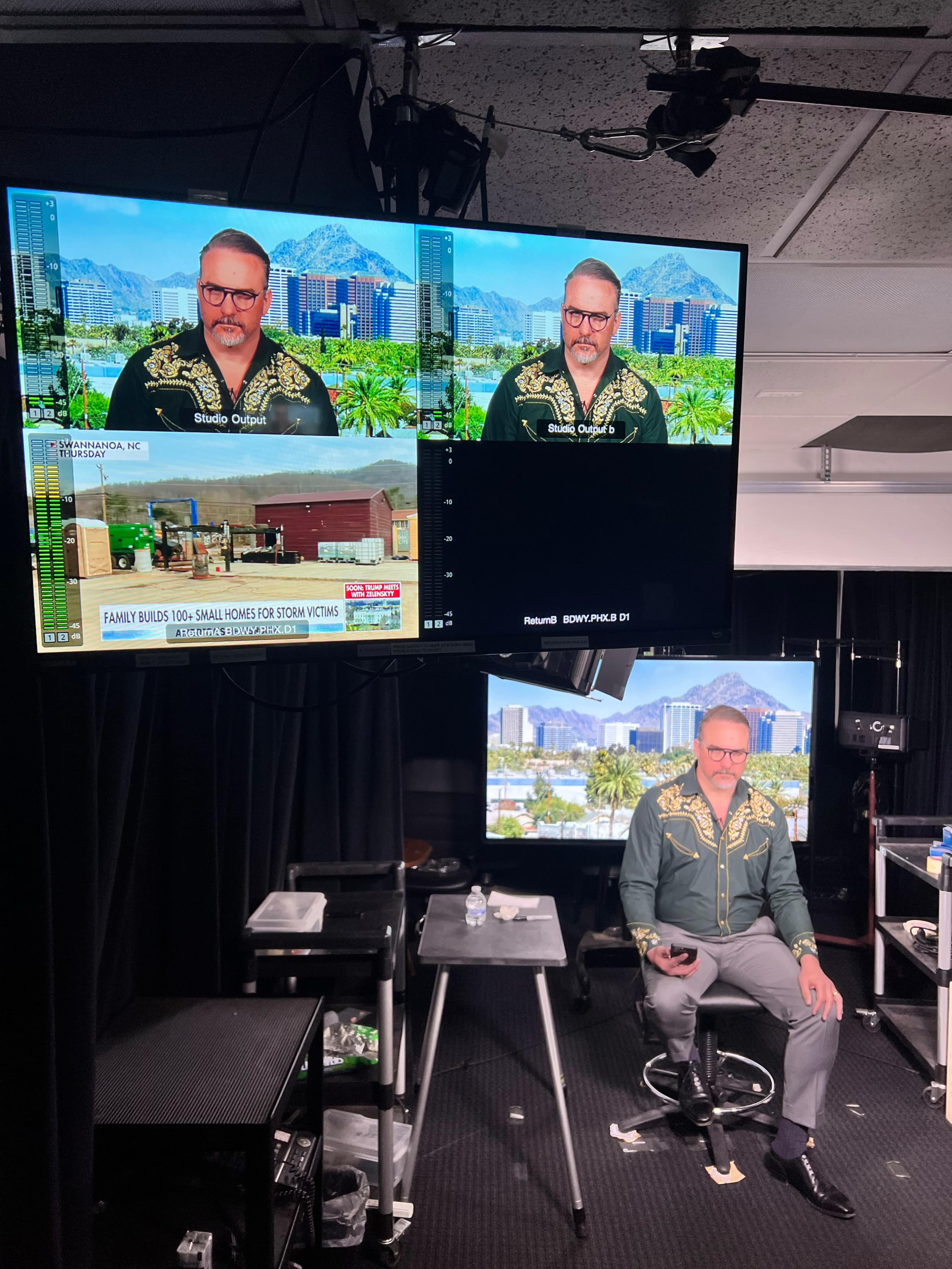 A man in a decorative shirt is seated in a television studio with multiple screens displaying news content and a city skyline in the background.