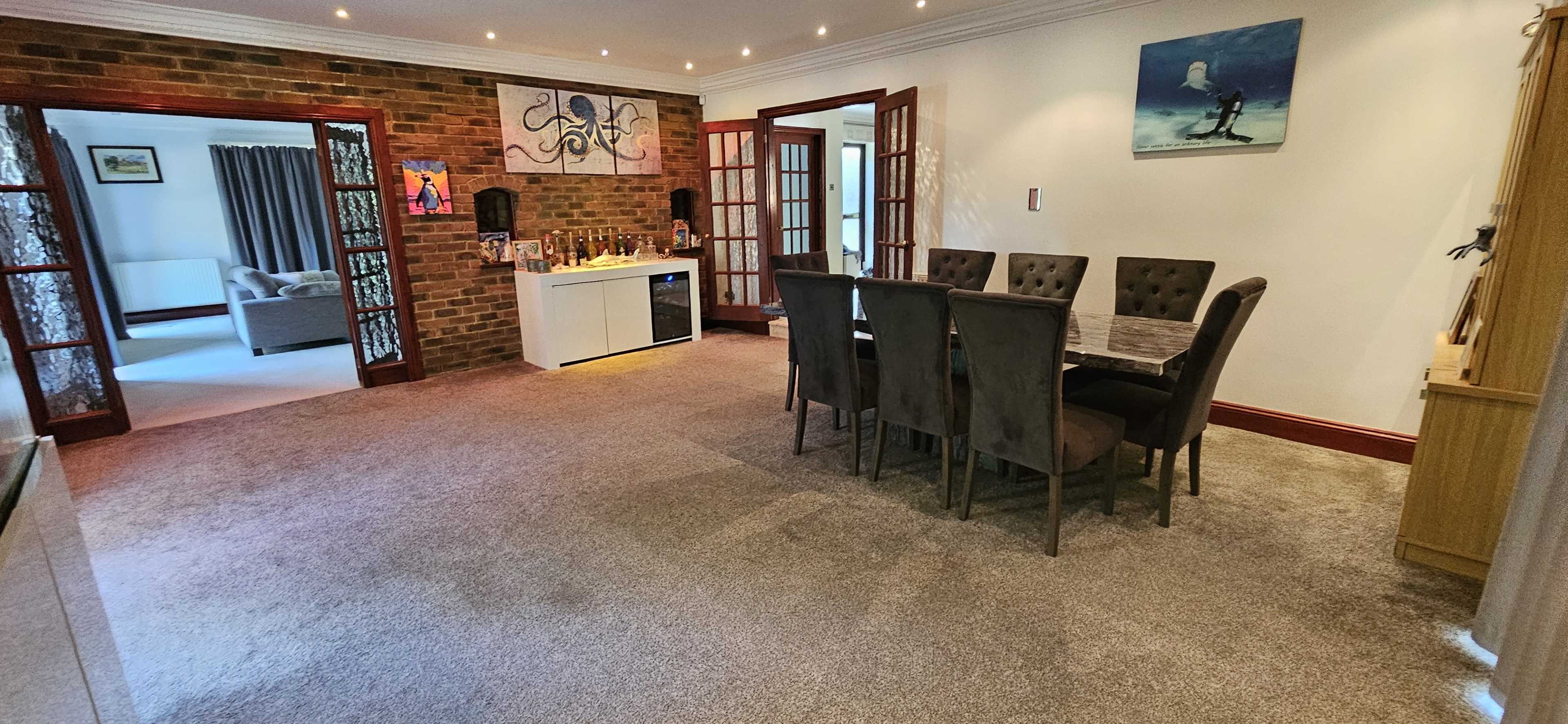 The image shows a spacious interior area with a dining table and chairs, adjacent to a bar setup, featuring brick walls and a carpeted floor.