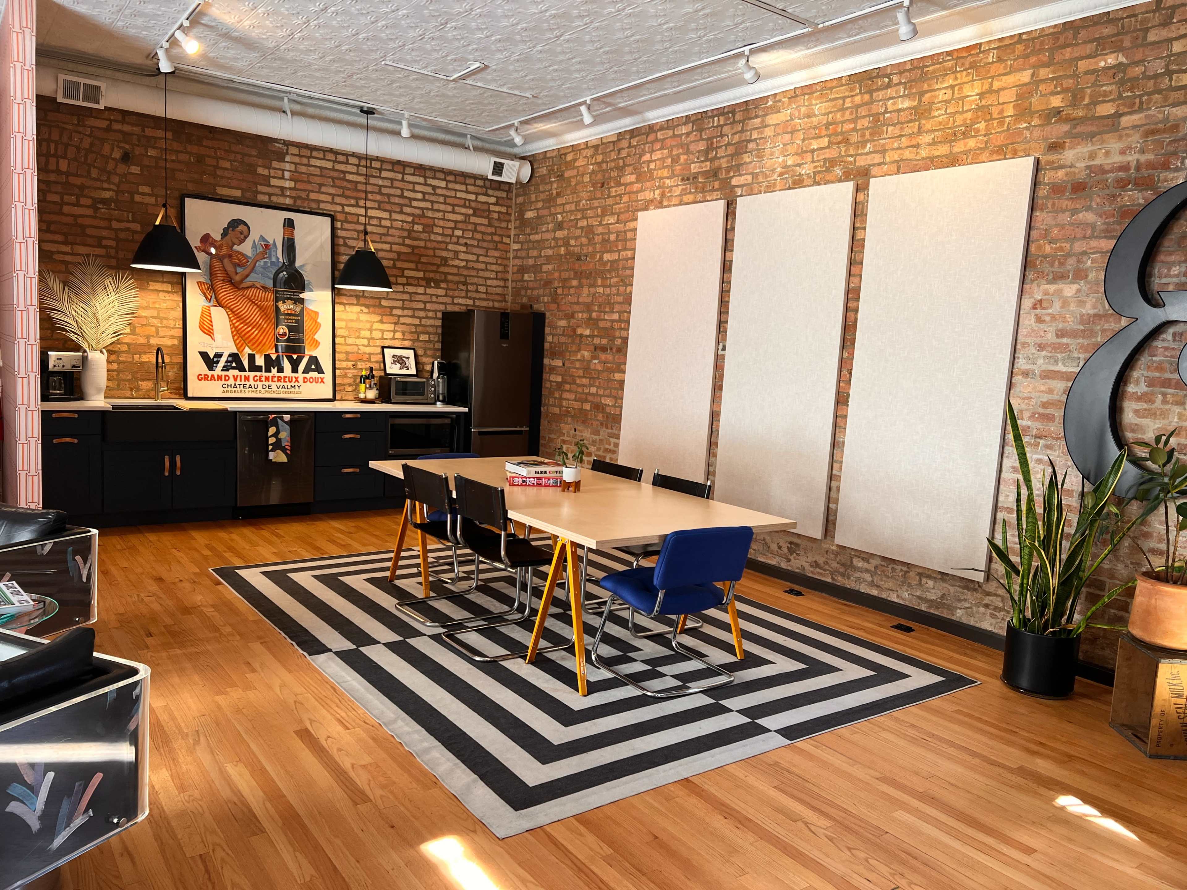 The image shows a modern interior with a dining table surrounded by chairs, a kitchenette in the background, and large, blank wall panels in a space with exposed brick walls and hardwood flooring.