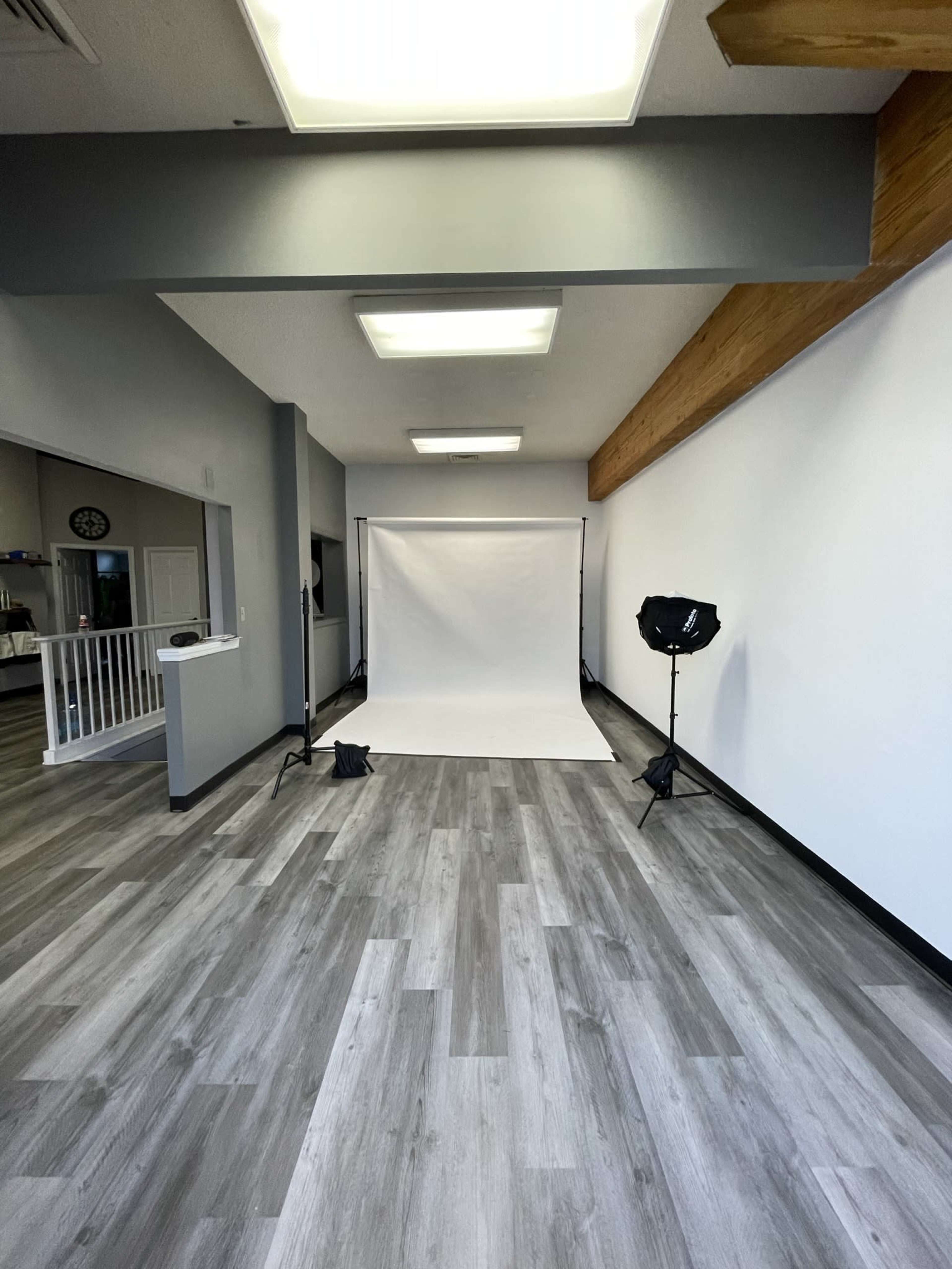 A spacious photography studio with a plain white backdrop and studio lighting set up against a light-colored wooden ceiling and gray flooring.