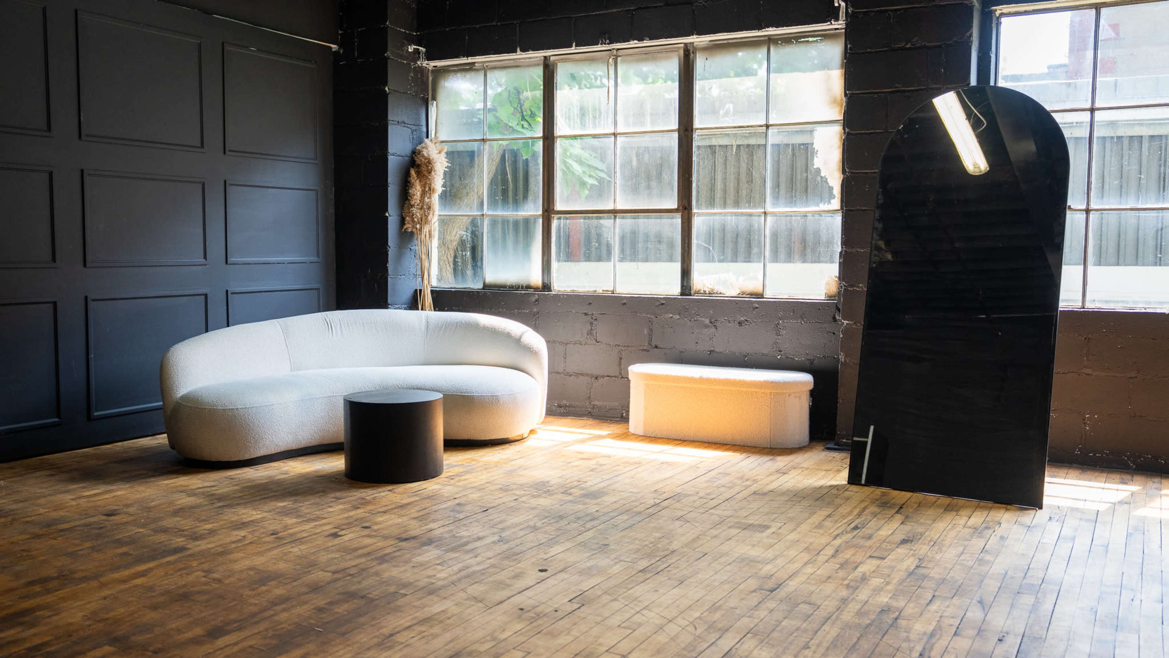 The image shows a modern, minimalist interior with a curved gray sofa, a black circular coffee table, and a light-colored bench, all positioned near large windows in a room with wooden flooring.