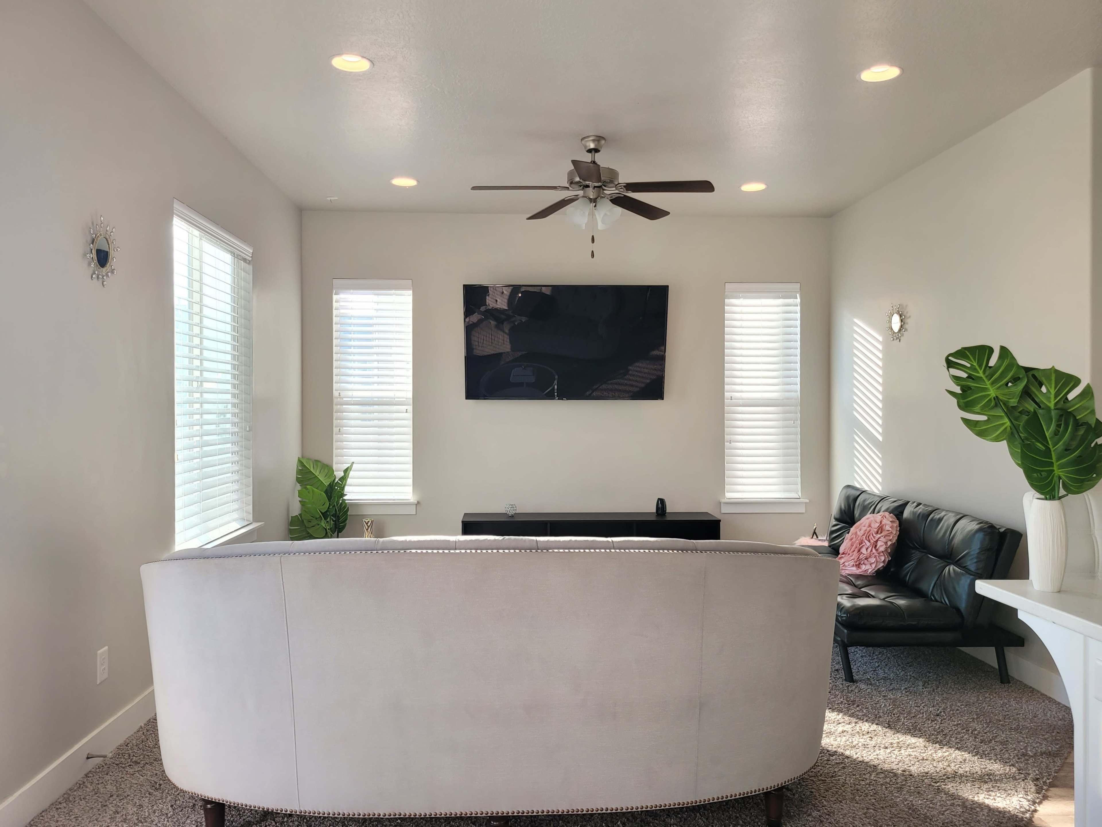 A modern living room with a light-colored sofa, a wall-mounted television, two windows, and a decorative plant.