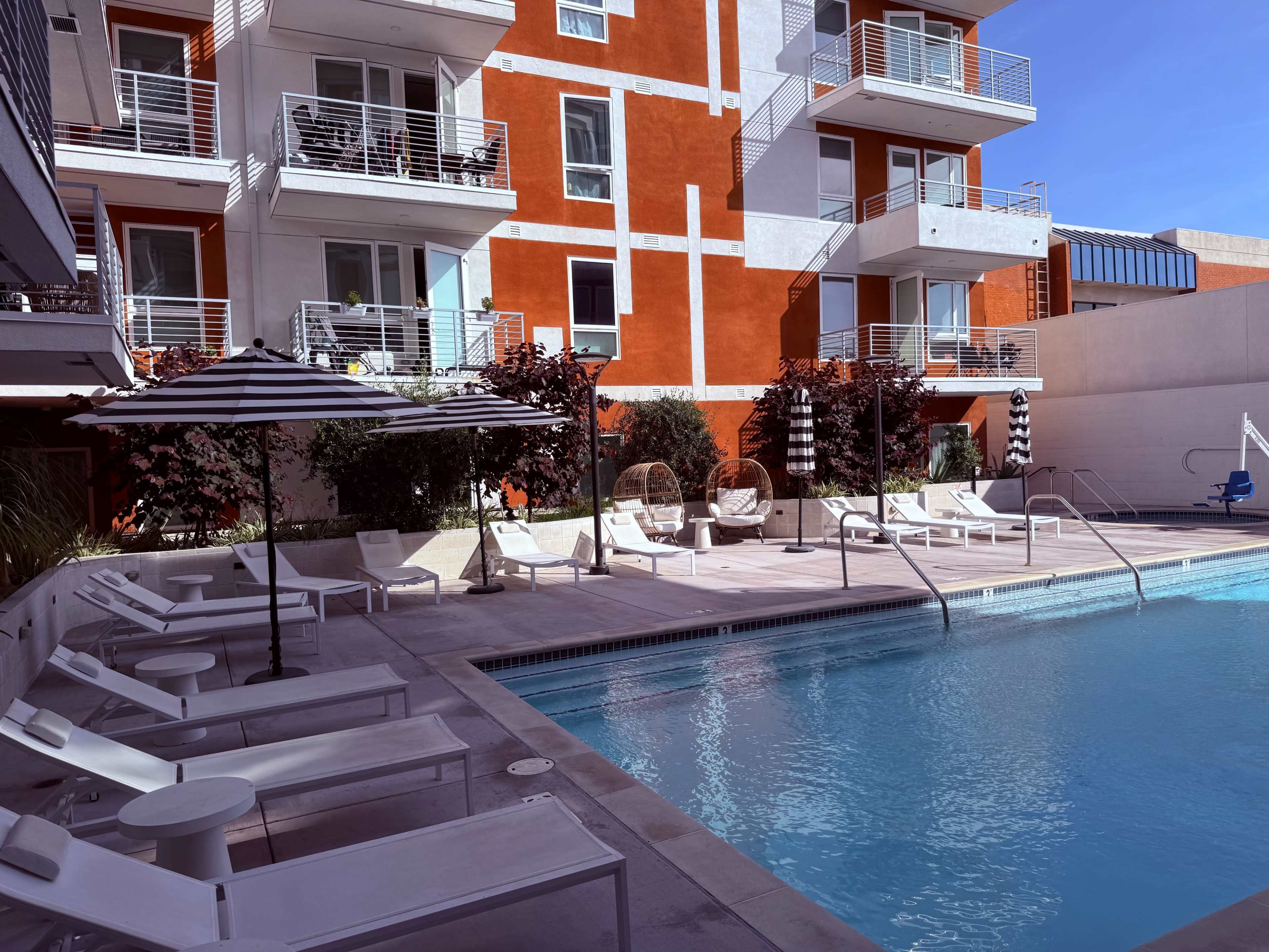 The image shows a modern outdoor pool area surrounded by lounge chairs, a striped umbrella, and colorful apartment buildings in the background.