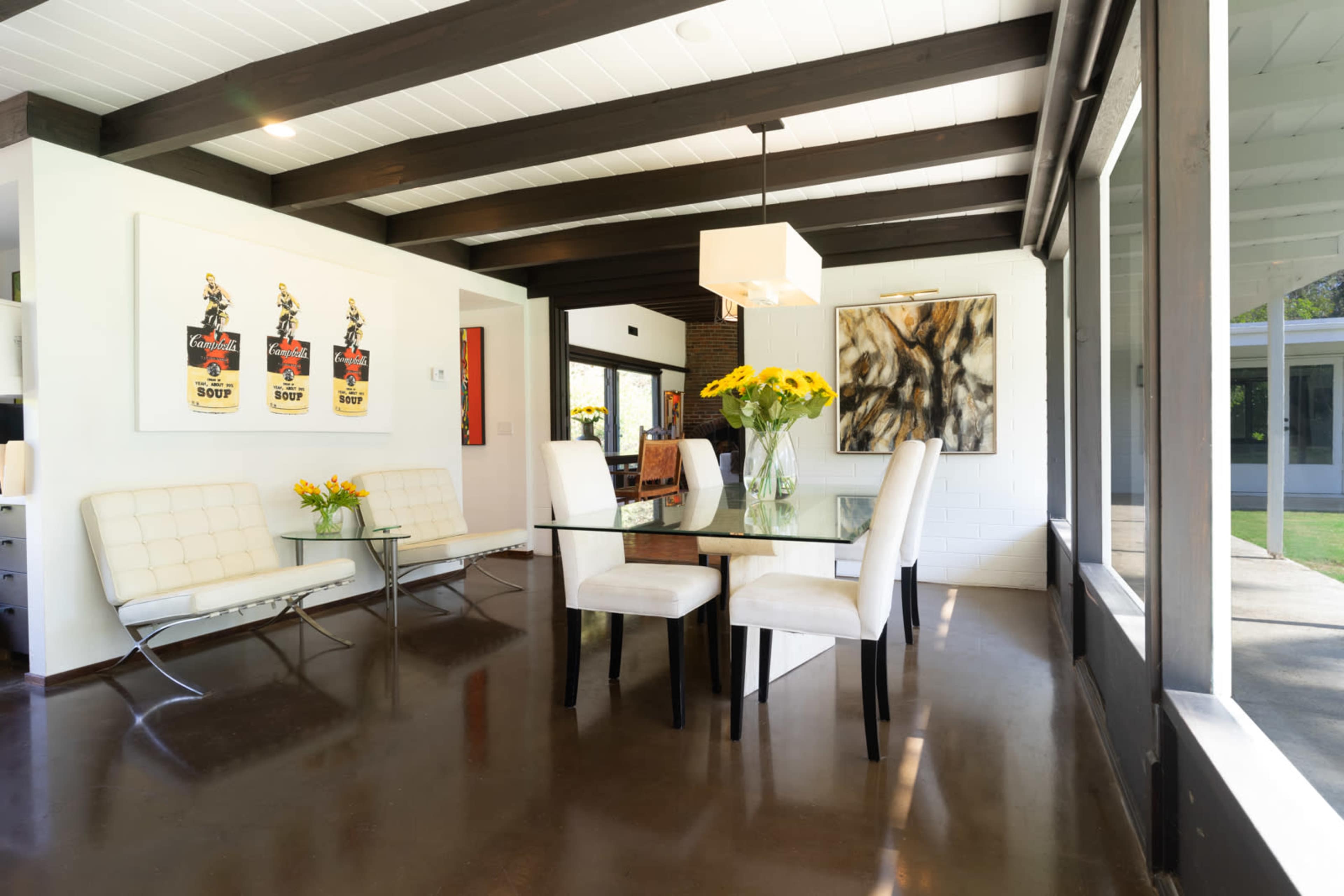 A modern dining area features a glass table surrounded by white upholstered chairs, with artwork on the walls and large windows allowing natural light.