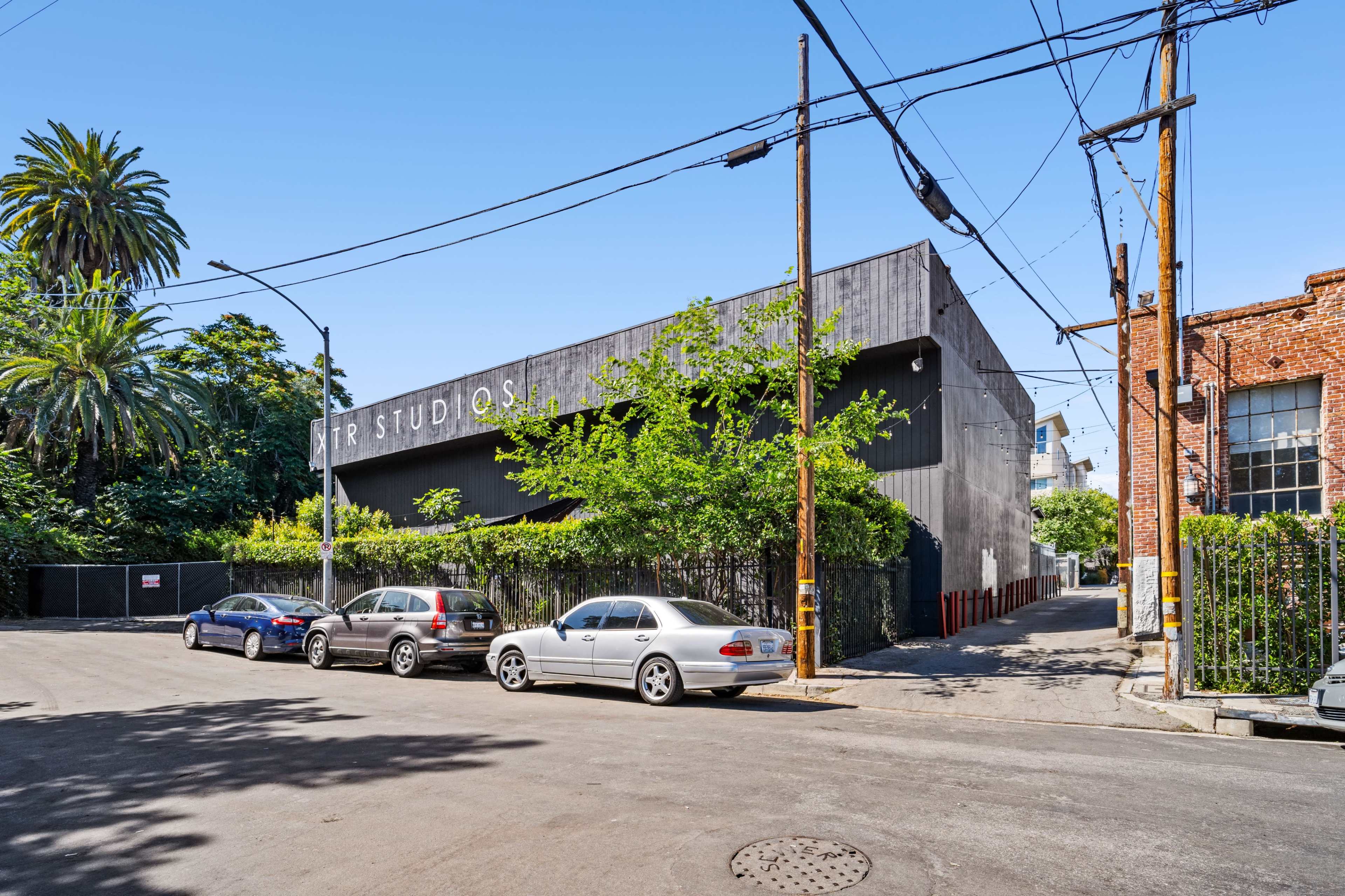 A large building with a sign labeled "ART STUDIOS" is situated at the corner of a street lined with parked cars and palm trees under a clear blue sky.
