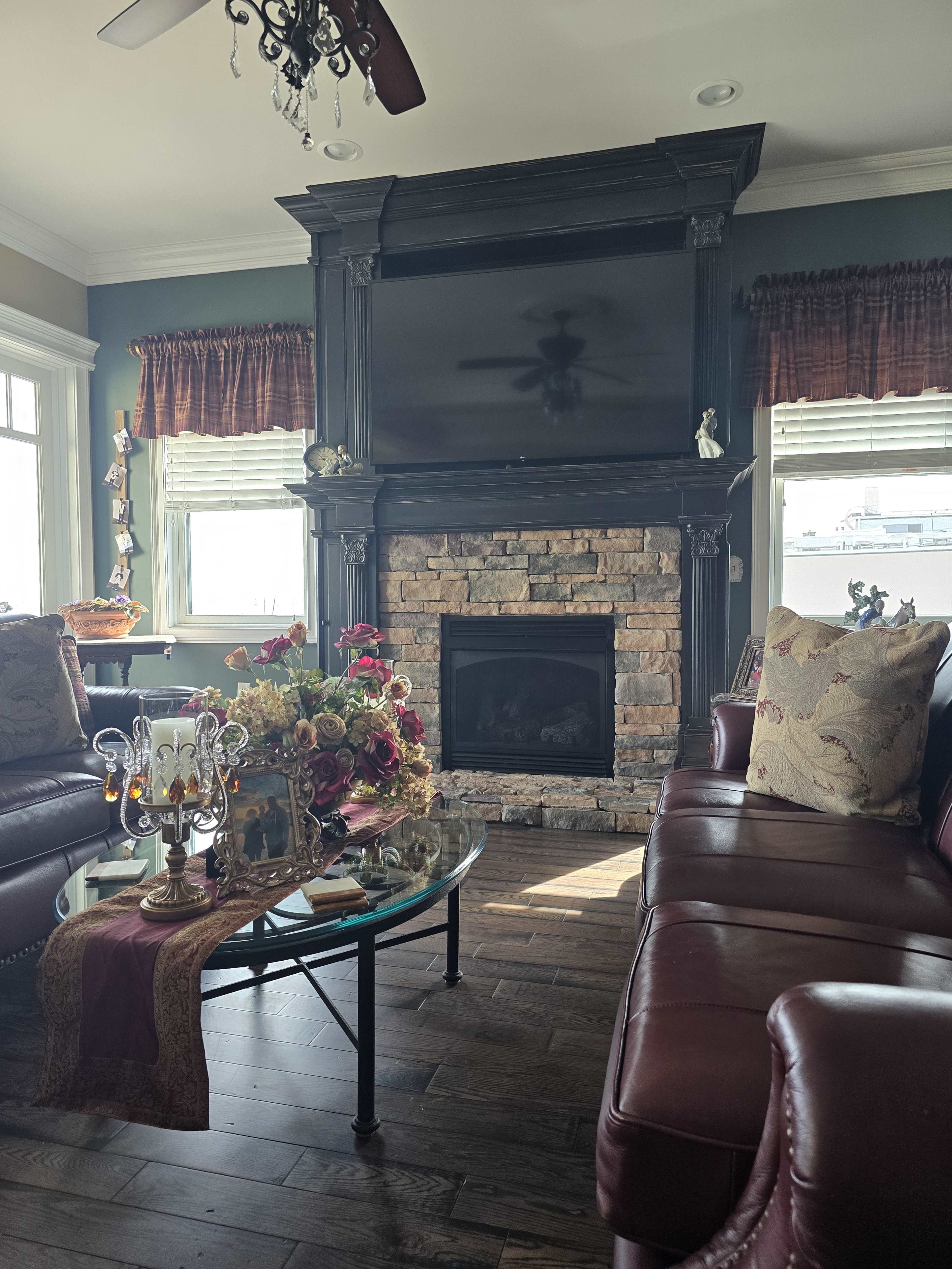 The living room features a stone fireplace with a large television above it, surrounded by leather sofas and a glass coffee table adorned with flowers.