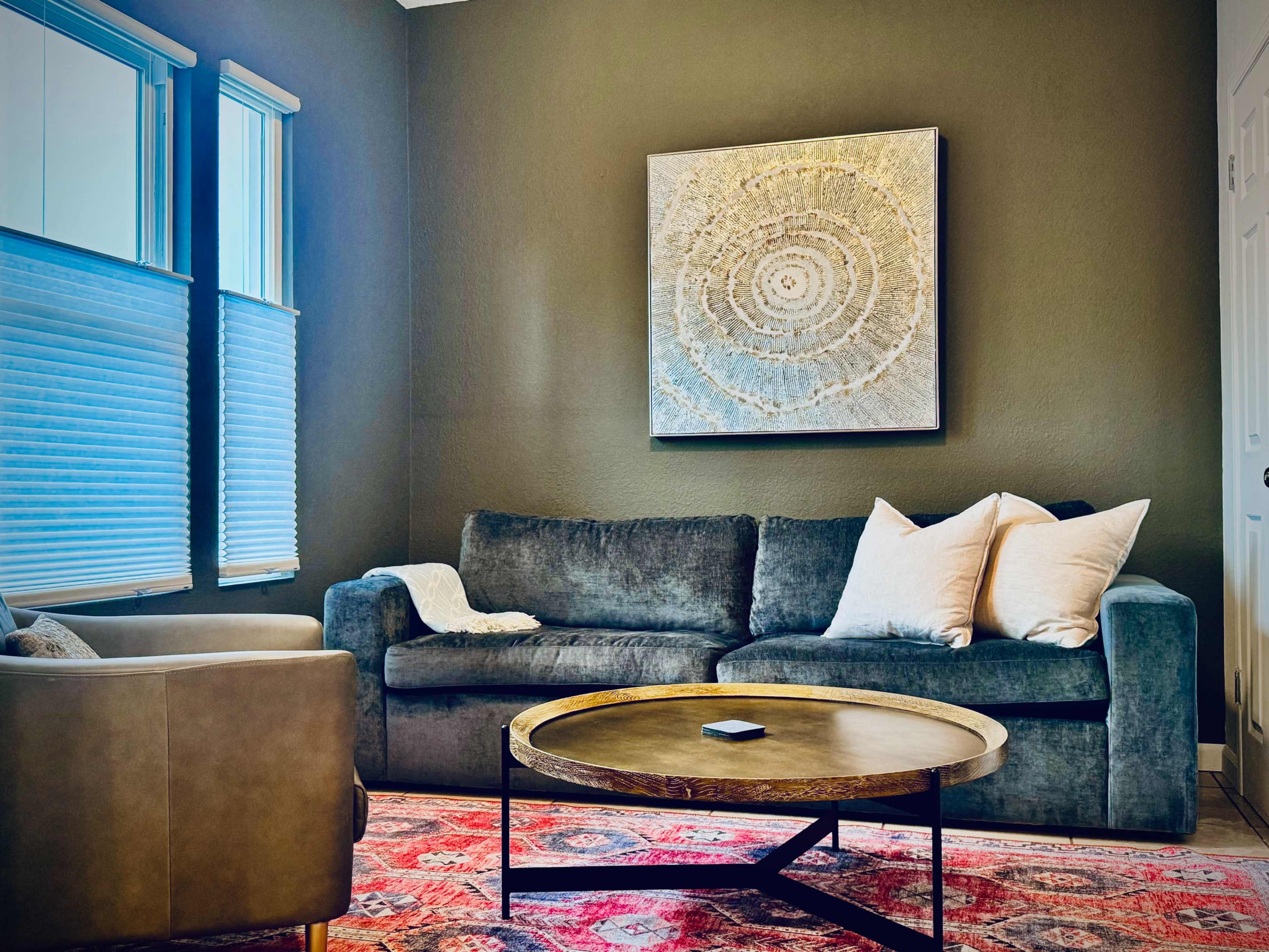 A living room features a gray sofa with white pillows, a round coffee table, and a large abstract painting on the wall.