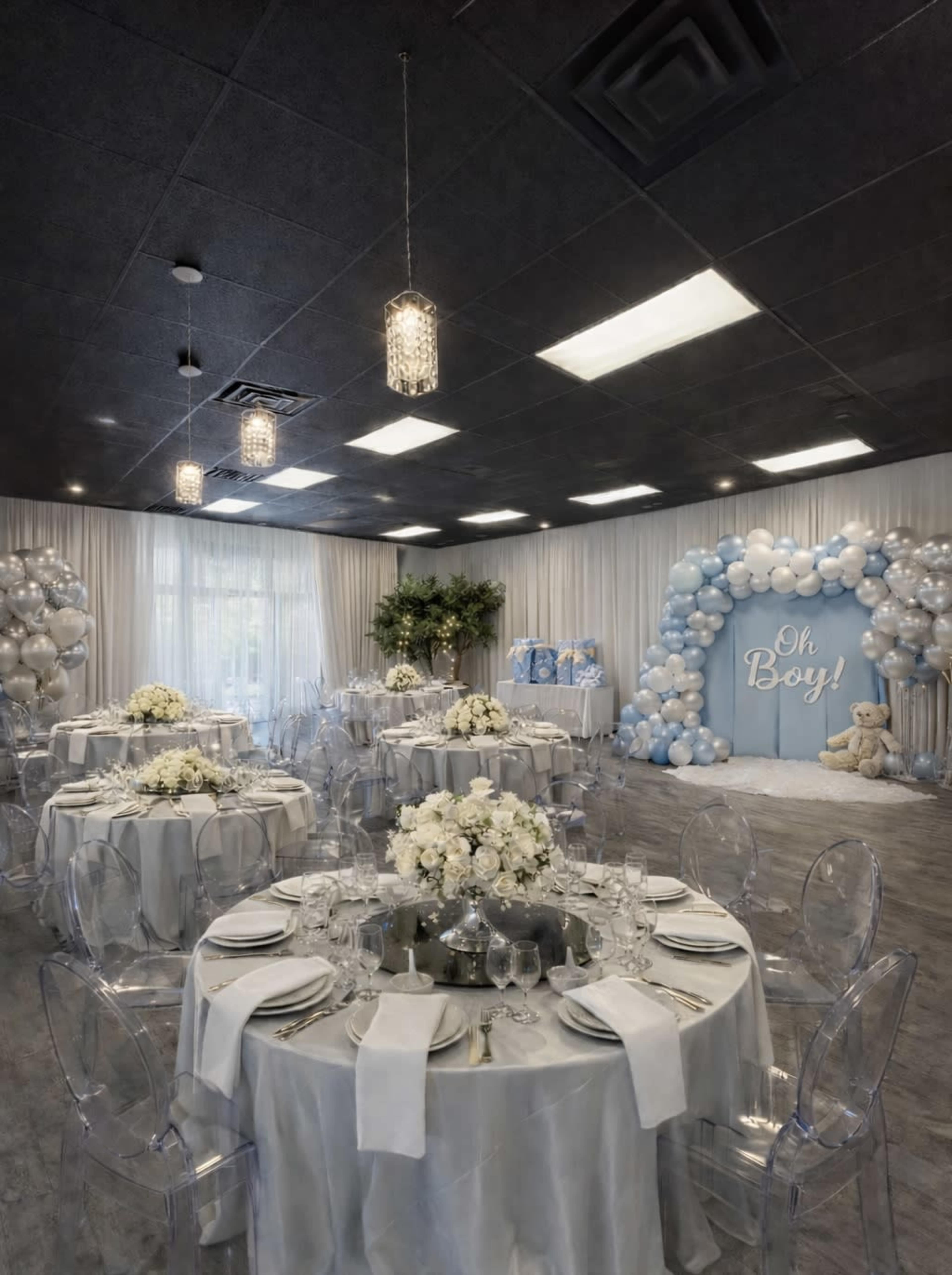 The image shows a decorated event space set up for a celebration, featuring round tables with floral centerpieces and a backdrop of blue and white balloons along with a sign that reads "Oh Boy!"