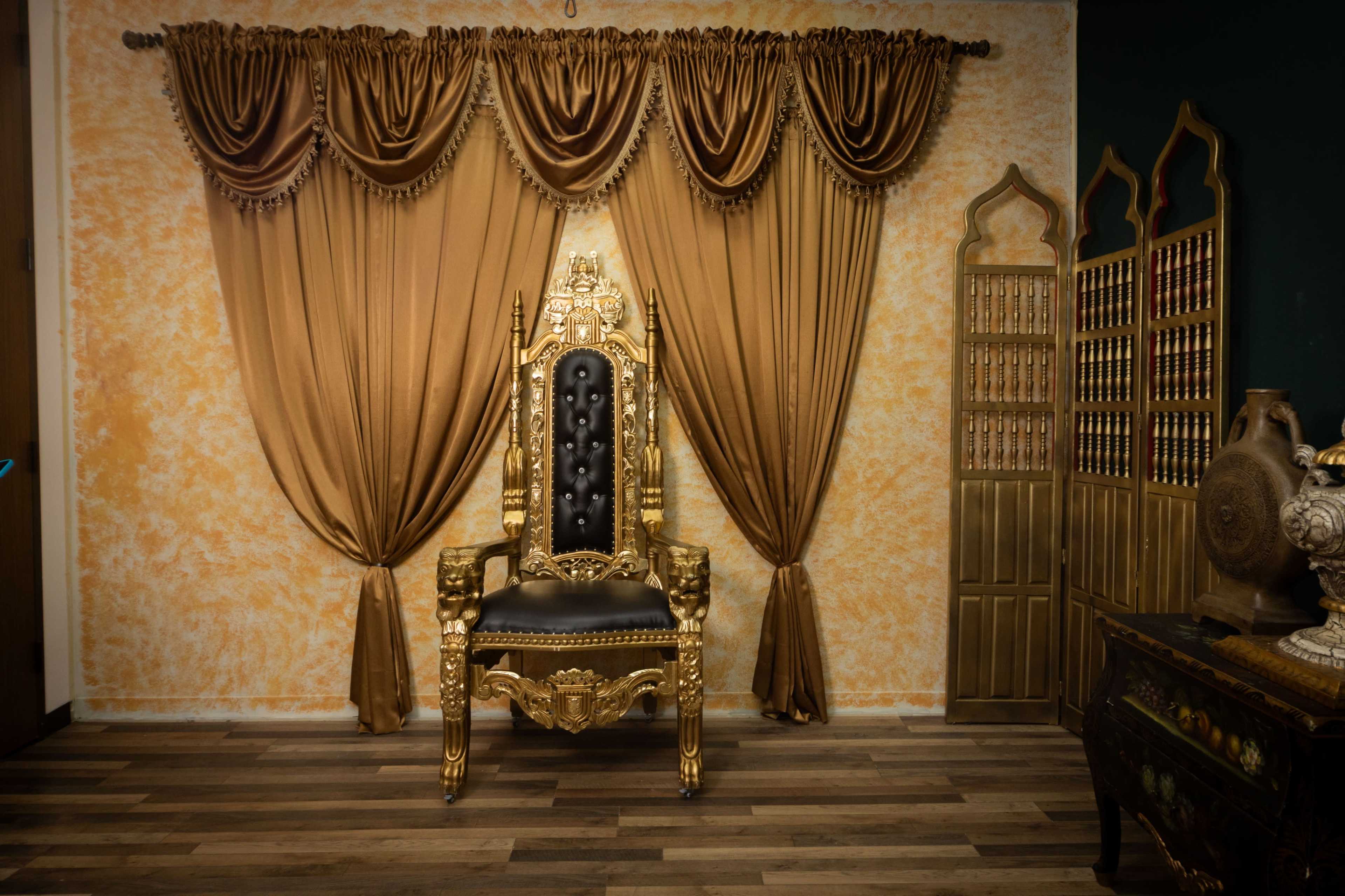 A gilded throne is positioned in front of elaborately draped golden curtains against a textured orange wall, complemented by decorative wooden furniture.