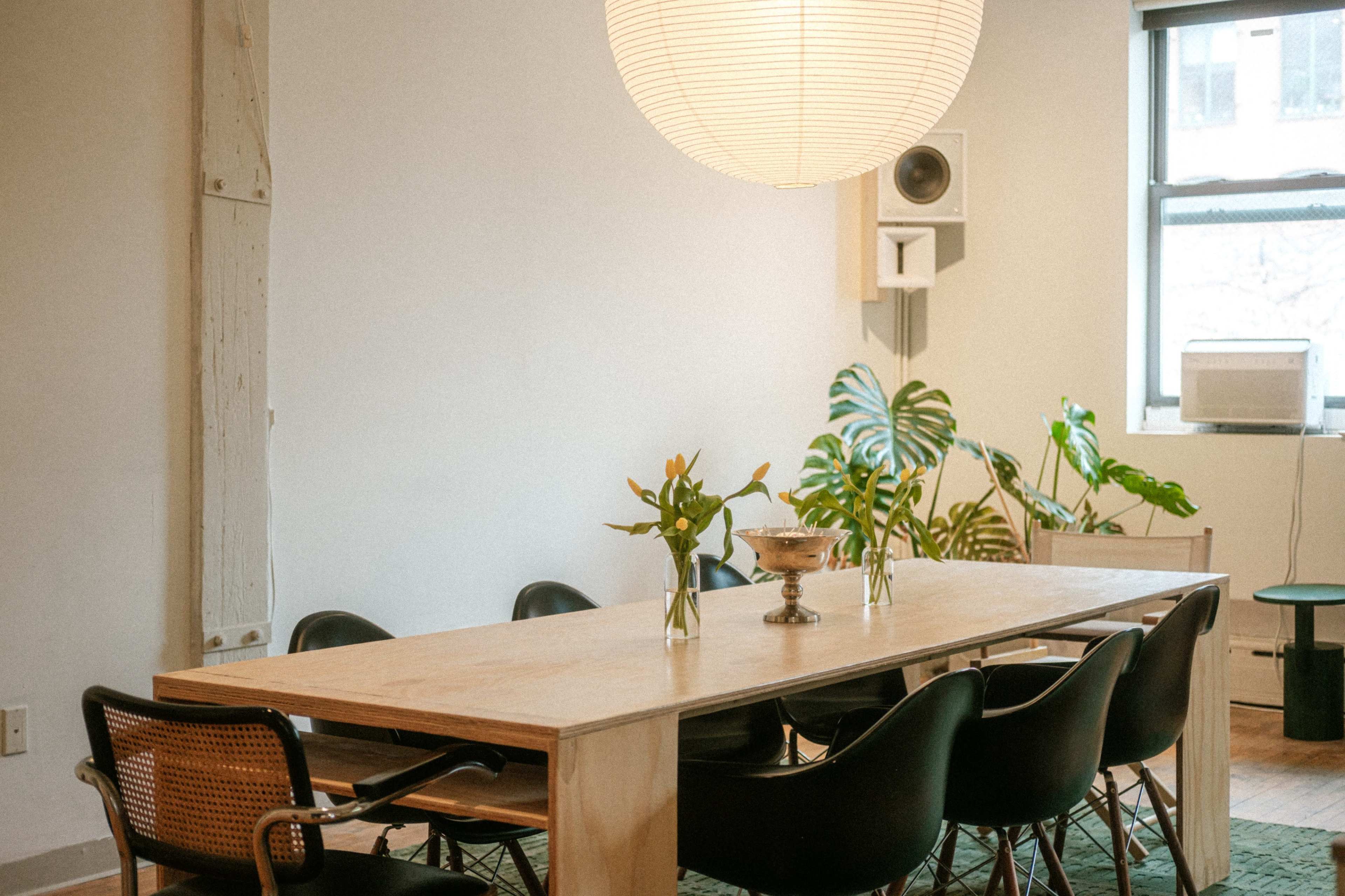 A dining area features a long wooden table surrounded by black chairs, with plants and a large pendant light overhead.