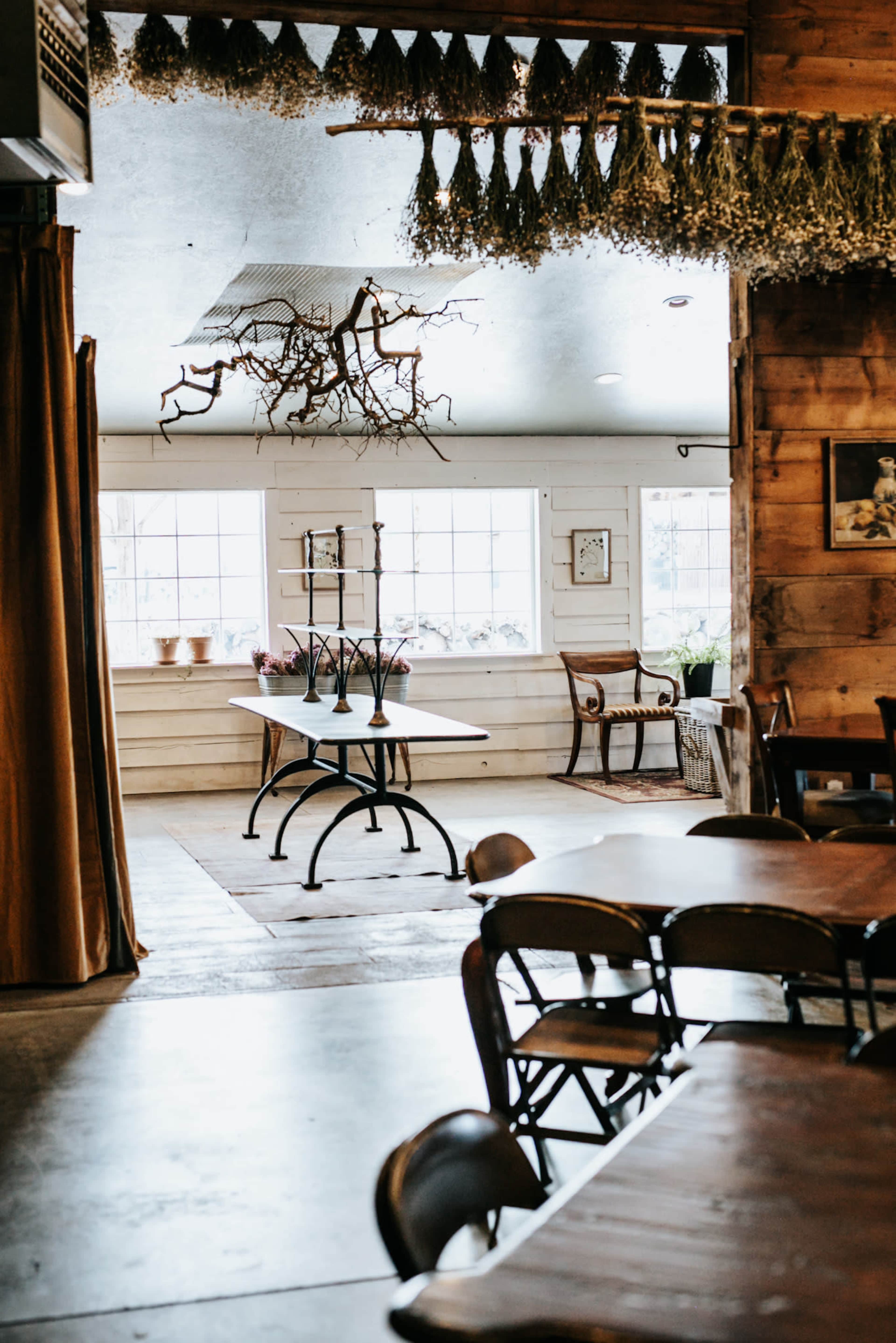 The image shows a rustic interior of a cozy dining space with wooden tables, a decorative ceiling, and large windows letting in natural light.