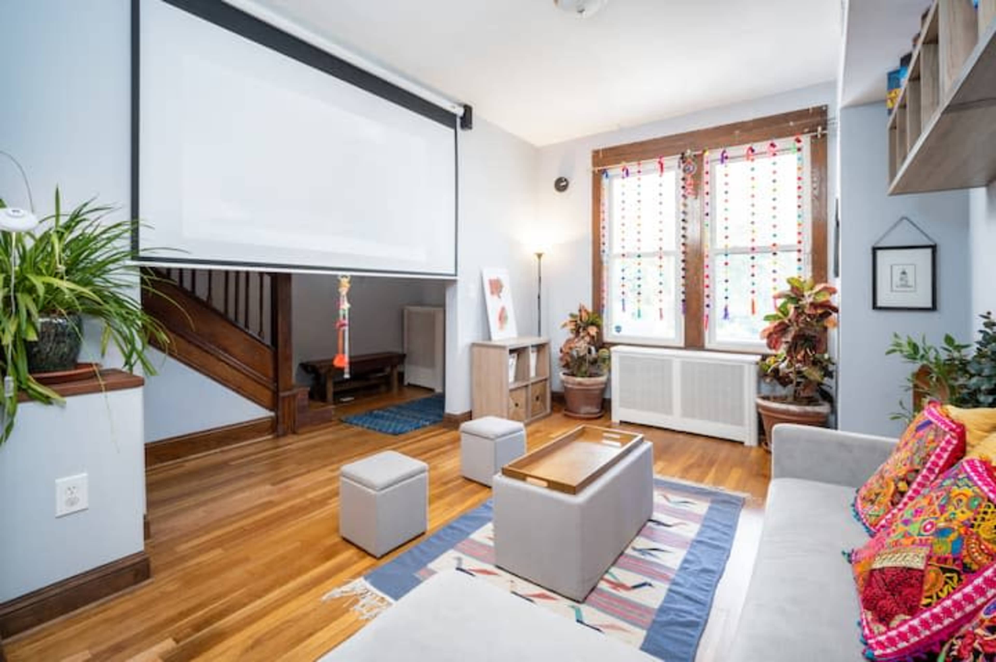 A living room with a projector screen, a gray sofa, colorful decorative cushions, plants, and a wooden staircase in the background.