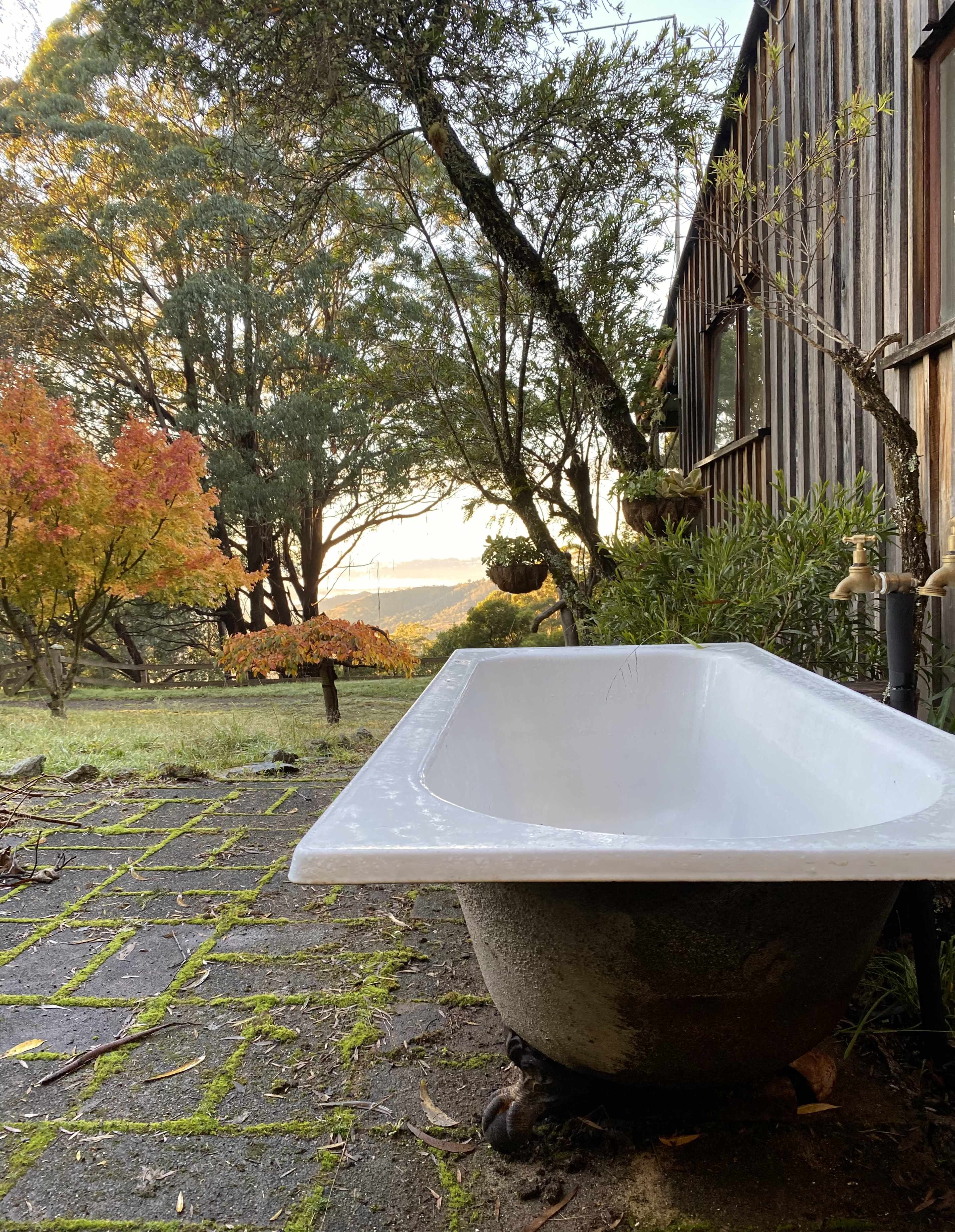 A freestanding bathtub is positioned outside on stone tiles, surrounded by greenery and autumn trees.