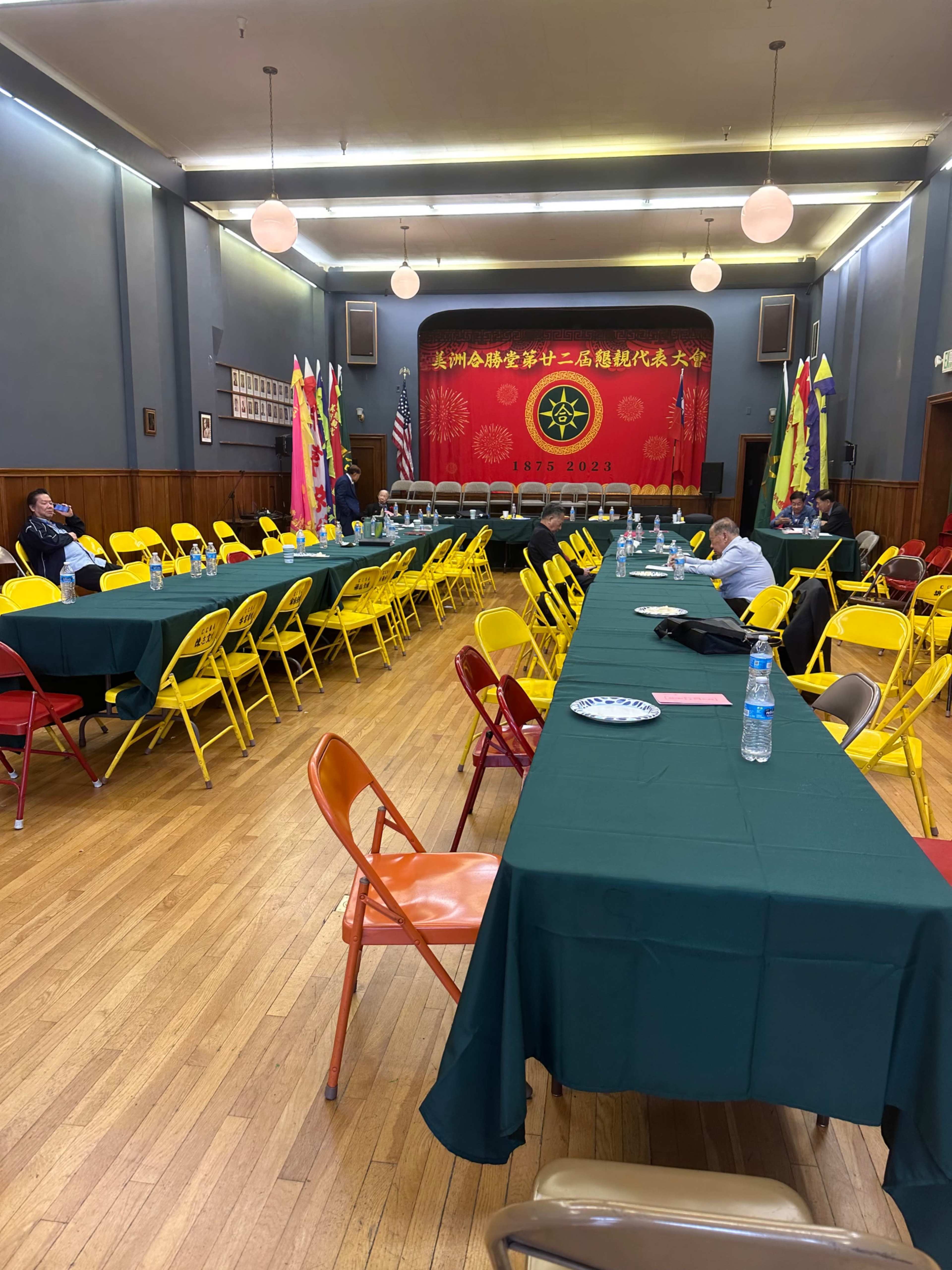 A spacious hall is set up with long tables covered in green tablecloths, surrounded by yellow and pink chairs, and decorated with flags and a large red banner.