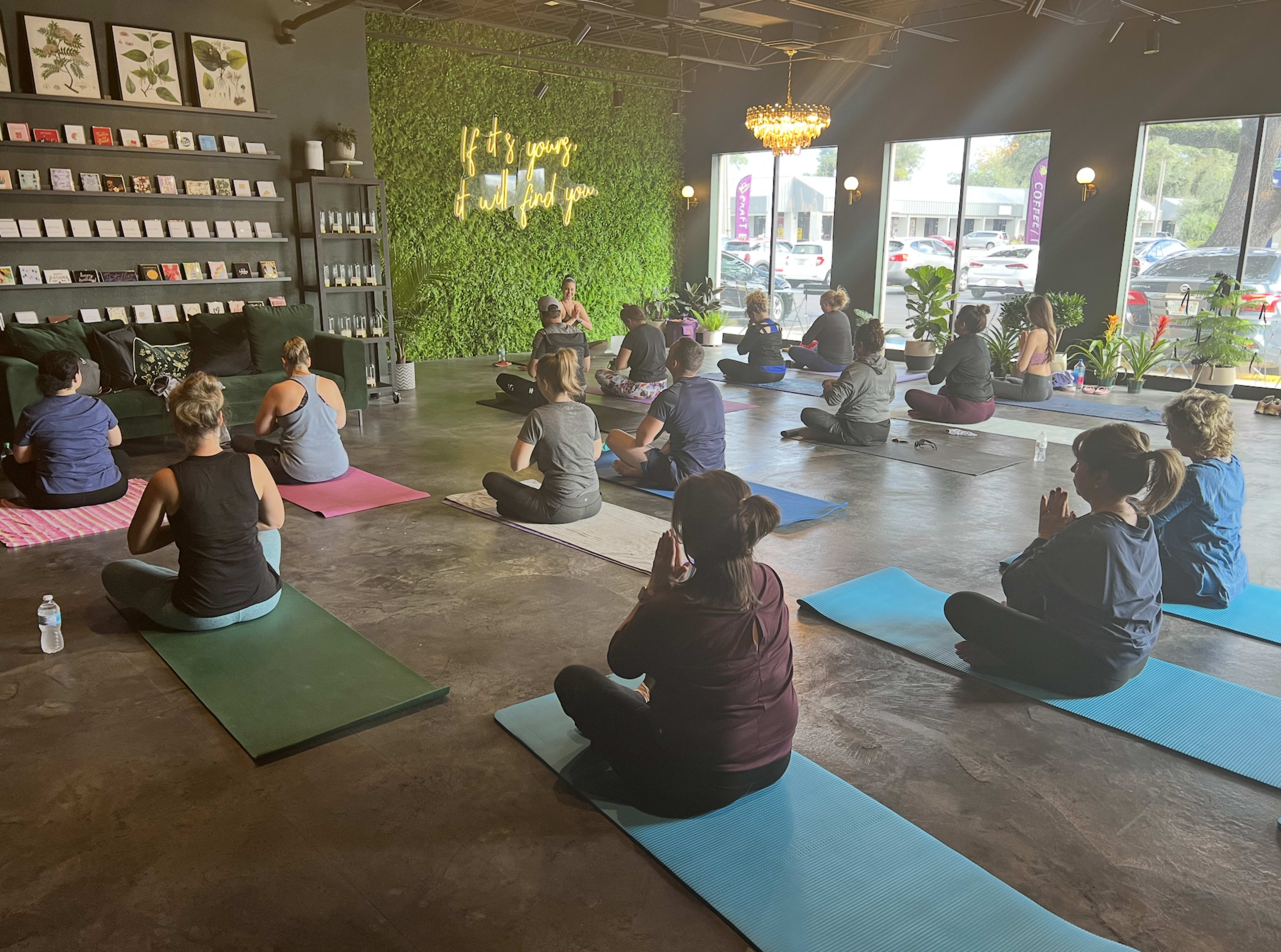 A group of people participates in a yoga class on mats inside a studio decorated with green plants and a large mural.