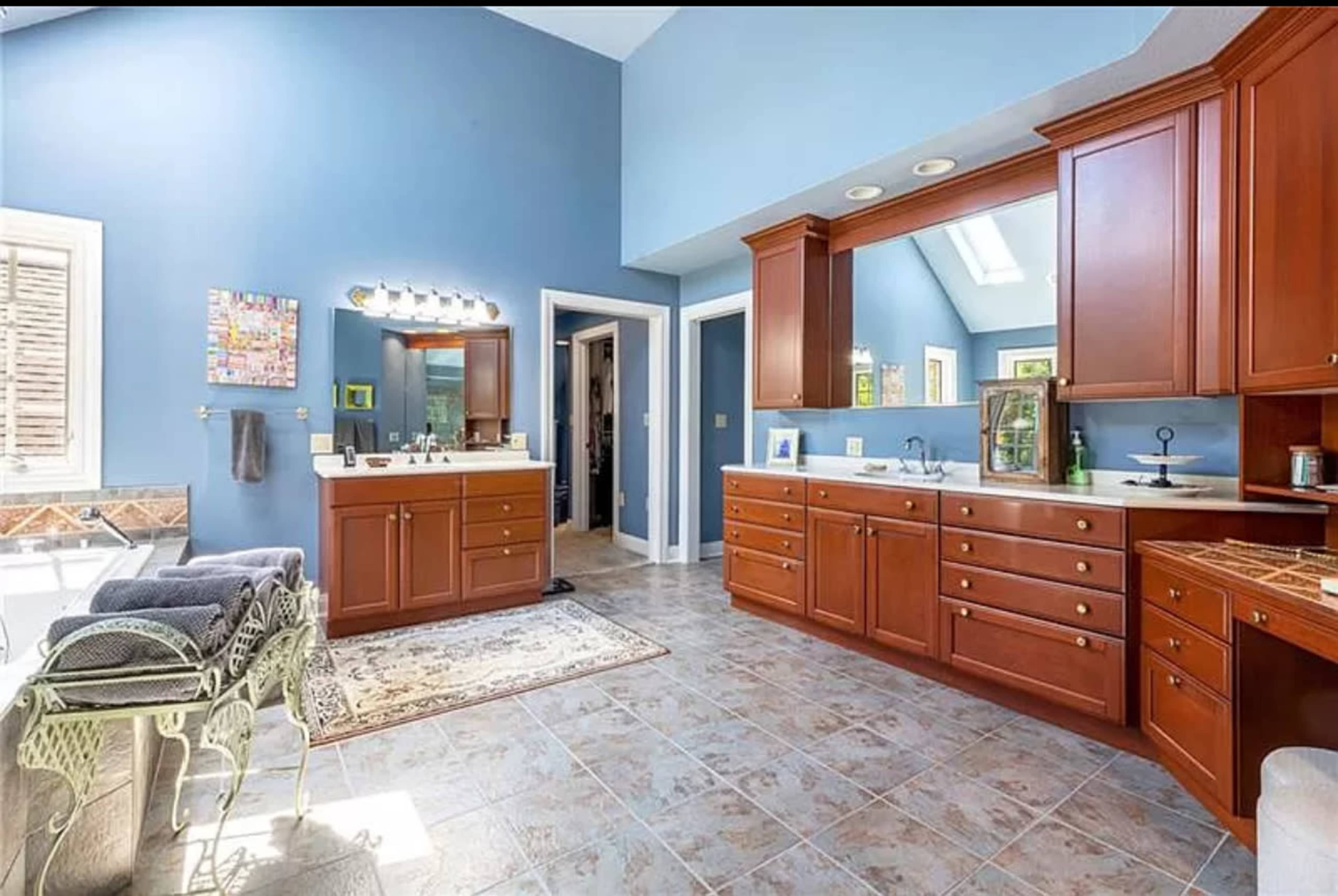 The image shows a spacious bathroom with blue walls, wooden cabinetry, and tiled flooring, featuring dual vanities and a seating area.