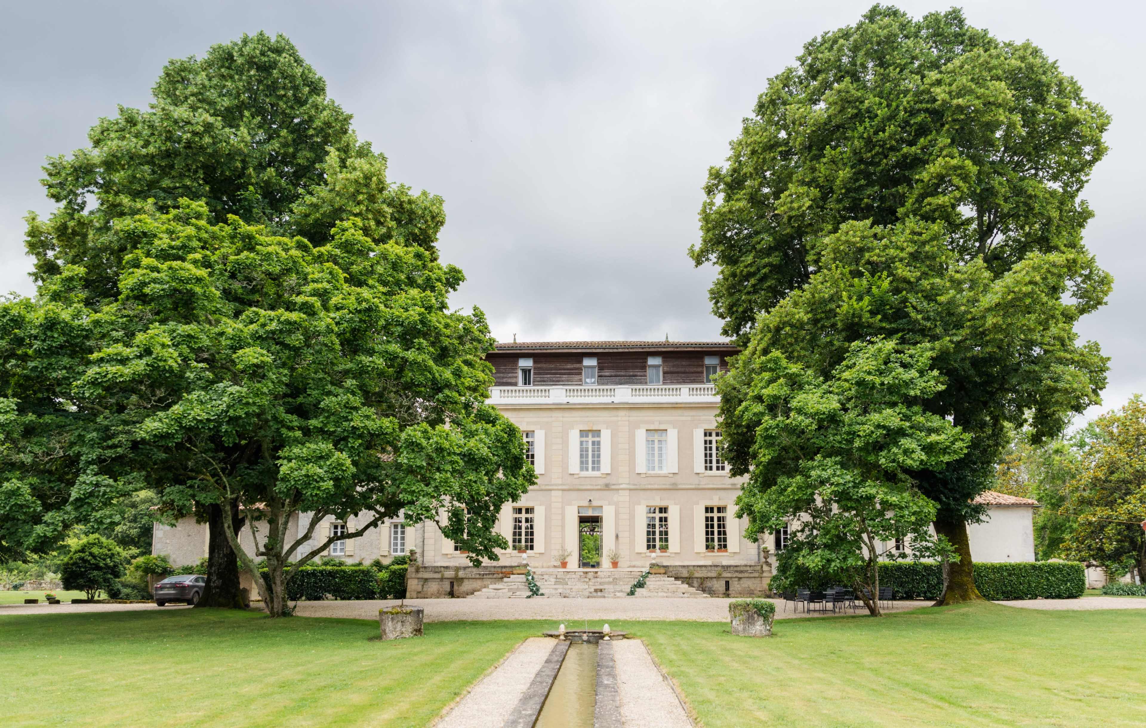 A large, two-story stone mansion is framed by tall trees in a landscaped garden.