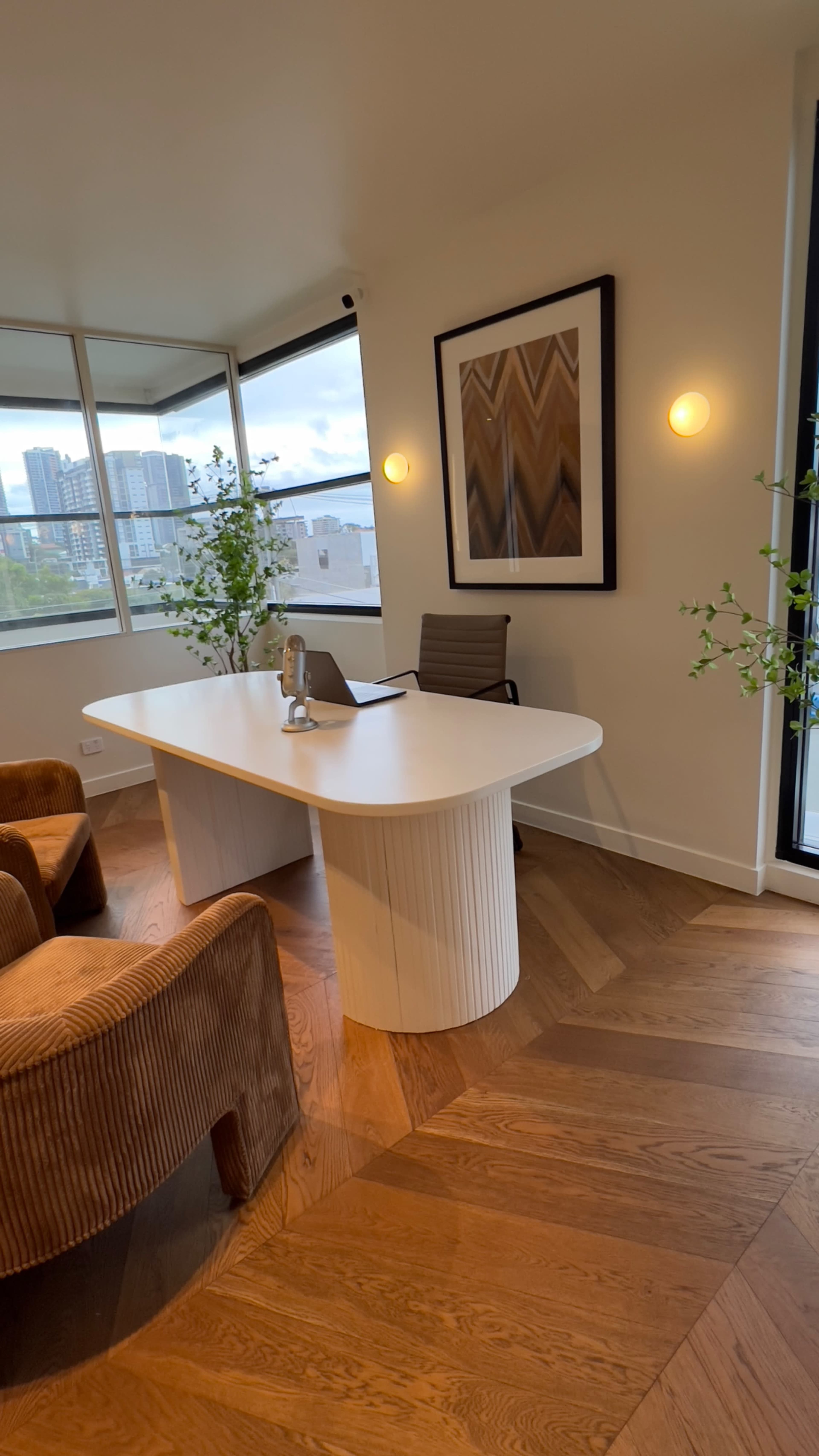 An office space features a curved white desk, a chair, and large windows overlooking a cityscape, with plants and modern decor.