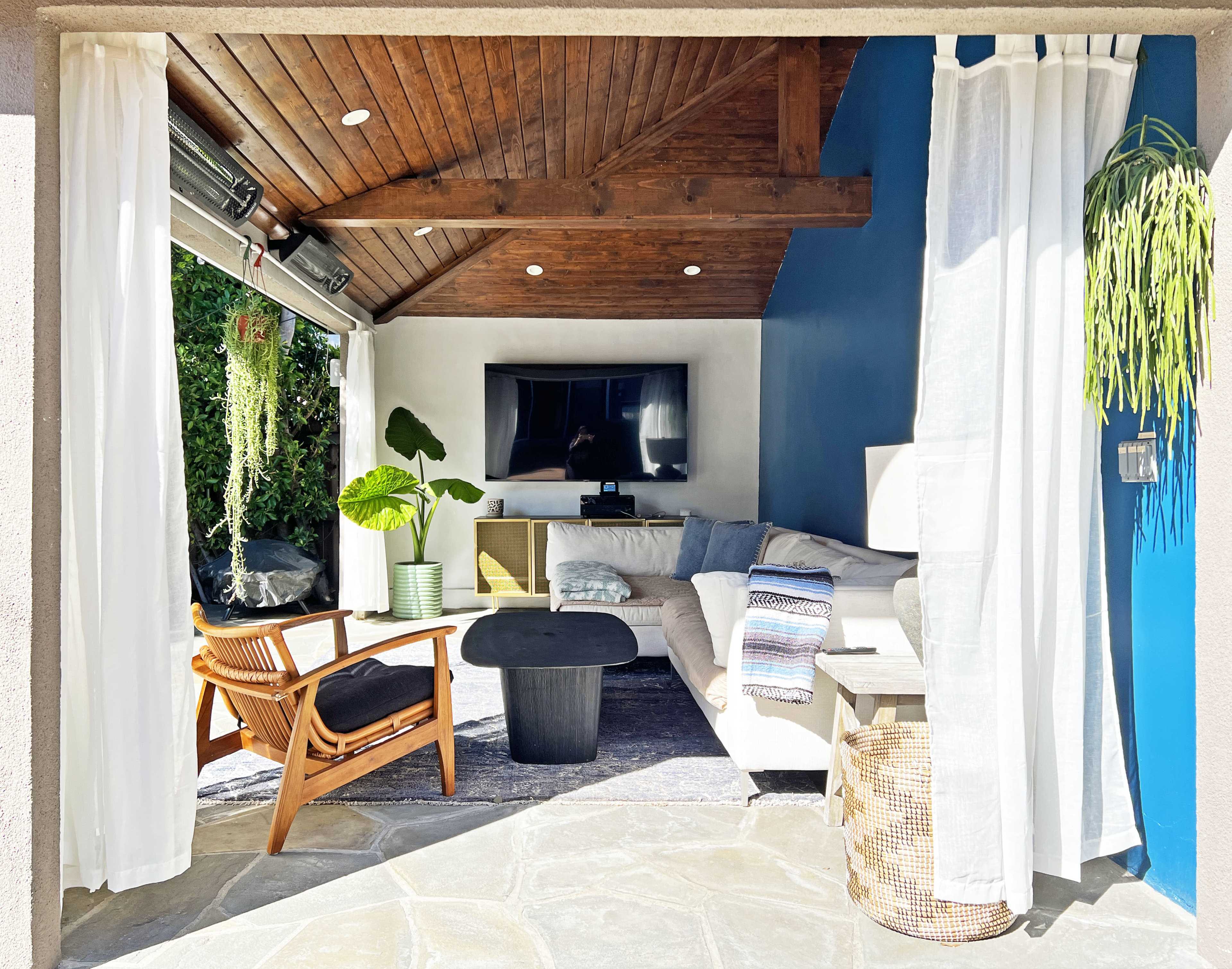 A modern living space with a sectional sofa, a television mounted on the wall, and large curtains framing an open doorway that leads to an outdoor area with greenery.