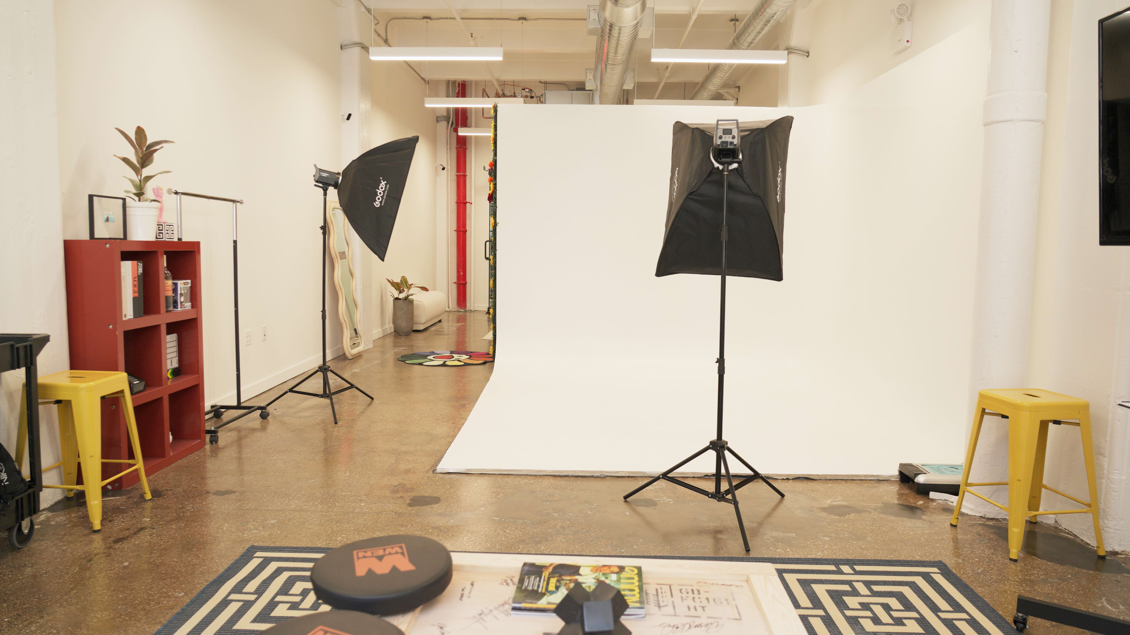 A photography studio with a white backdrop, two light stands, a colorful rug, and various props on a table.