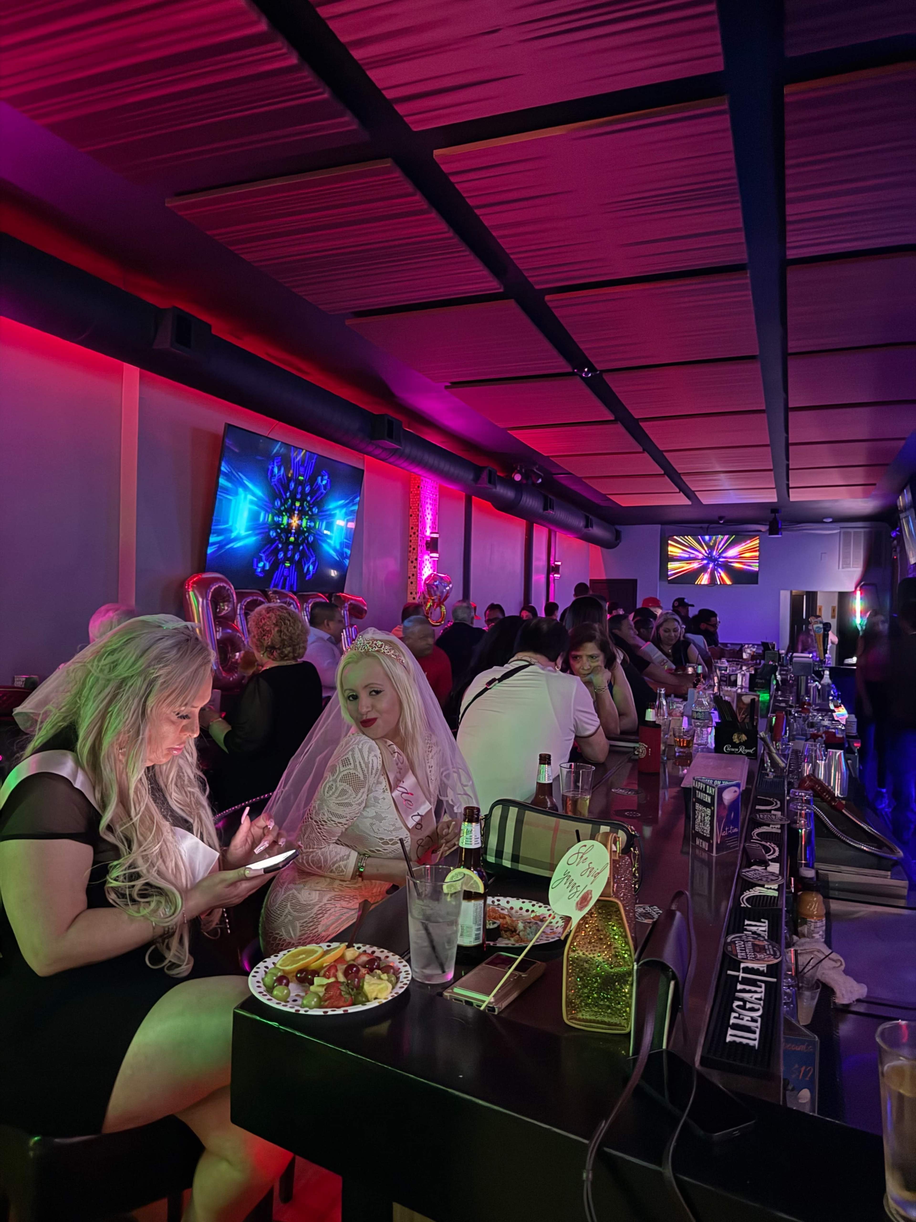 A bustling bar scene features patrons in formal attire, with one woman in a white dress and veil sitting at the counter, while colorful lights illuminate the space.