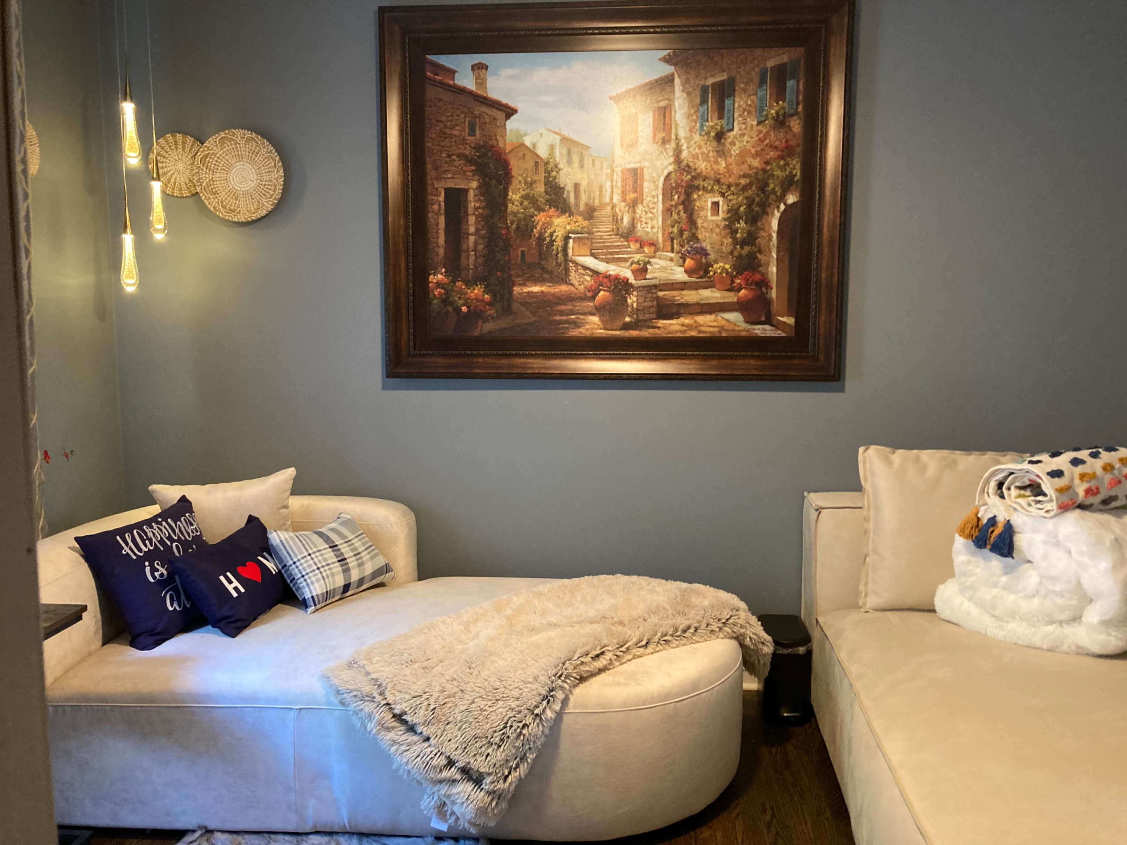 A cozy living room corner with a curved couch, decorative pillows, a fluffy throw, and a large painting of a scenic village as the focal point.
