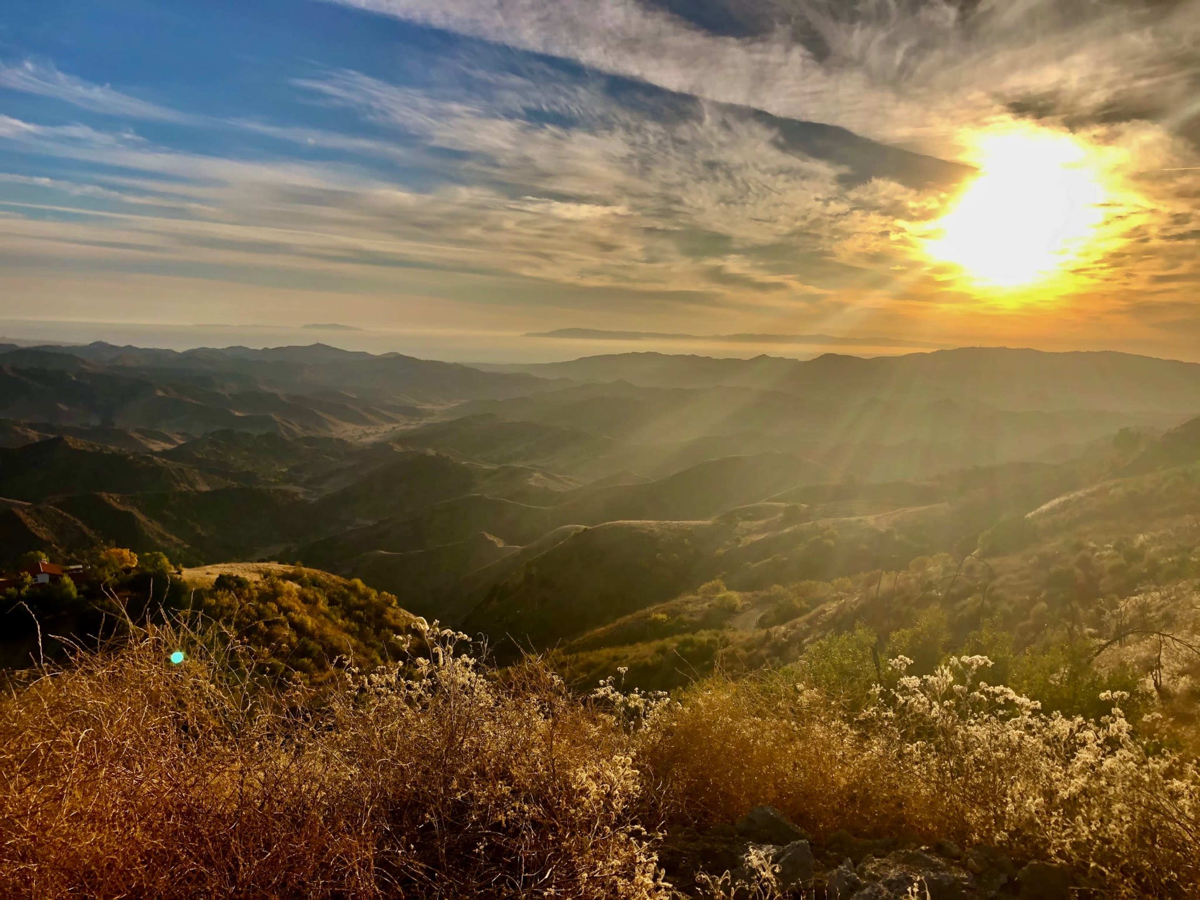 The sun sets over rolling hills, casting warm light on the landscape filled with dry grass and distant mountains.