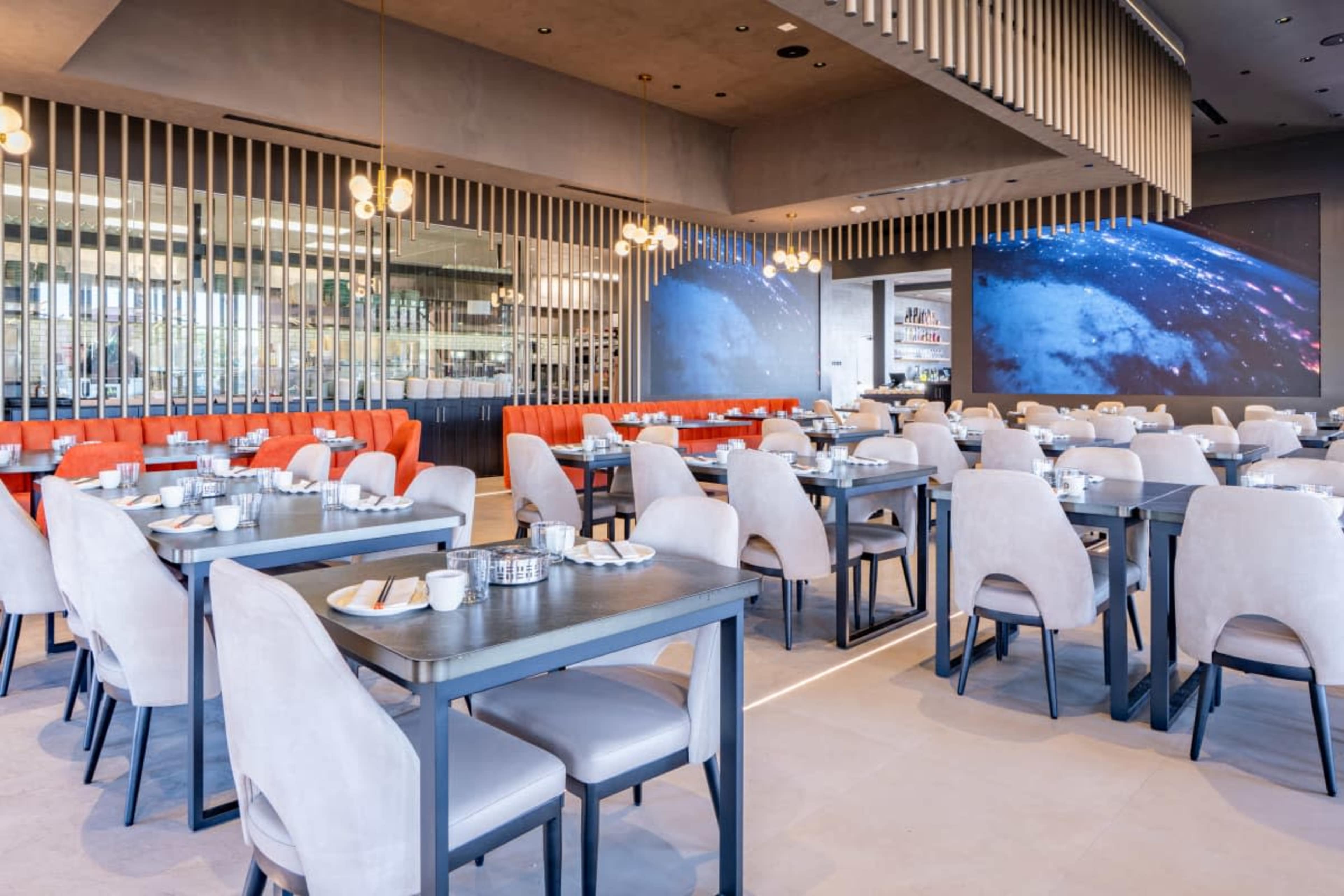 The image shows a modern restaurant interior with neatly arranged tables and chairs, large screens displaying visuals, and a bright atmosphere.