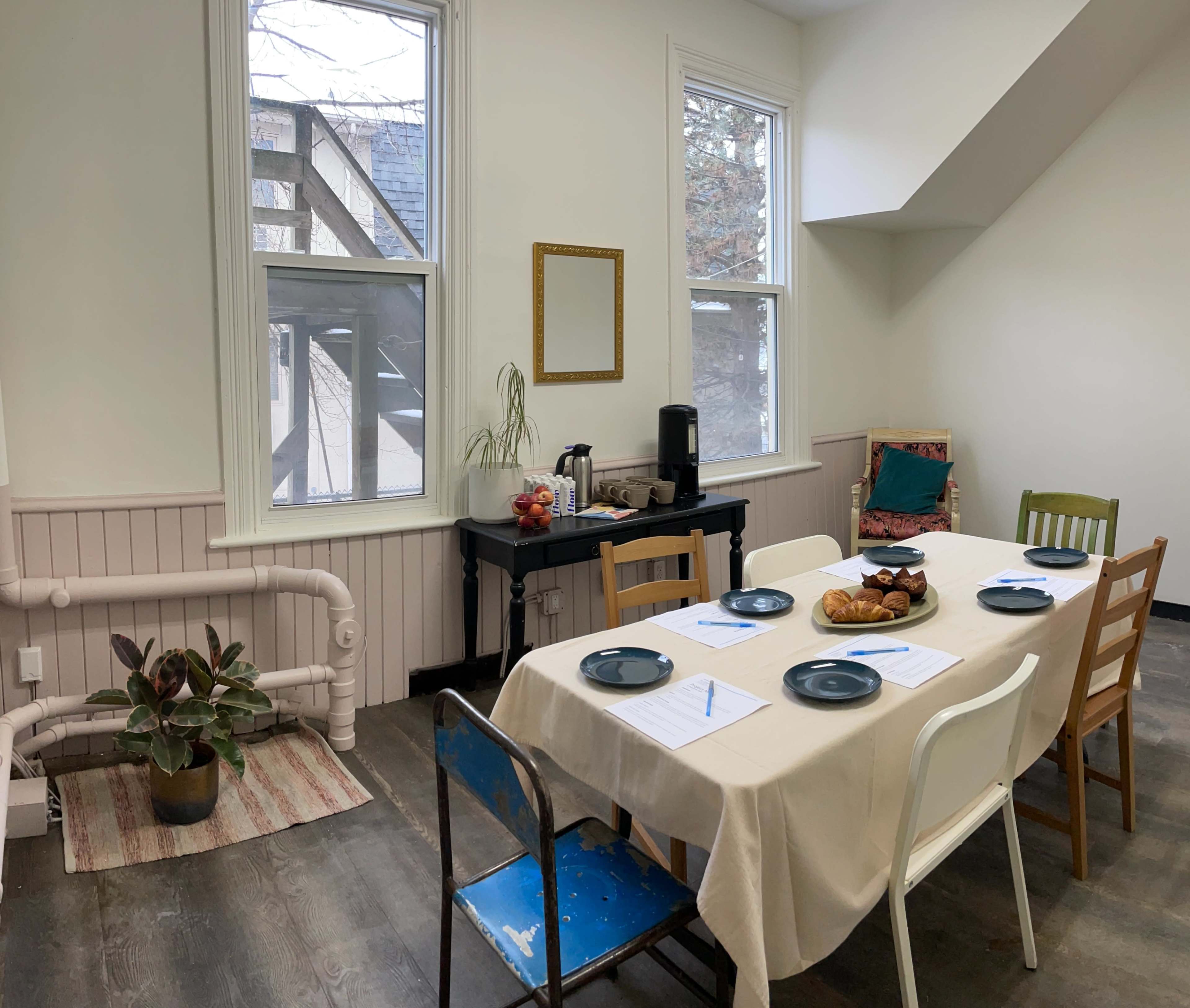 A dining area features a long table set with plates and a variety of food, surrounded by chairs of different colors and styles, with natural light coming through the windows.