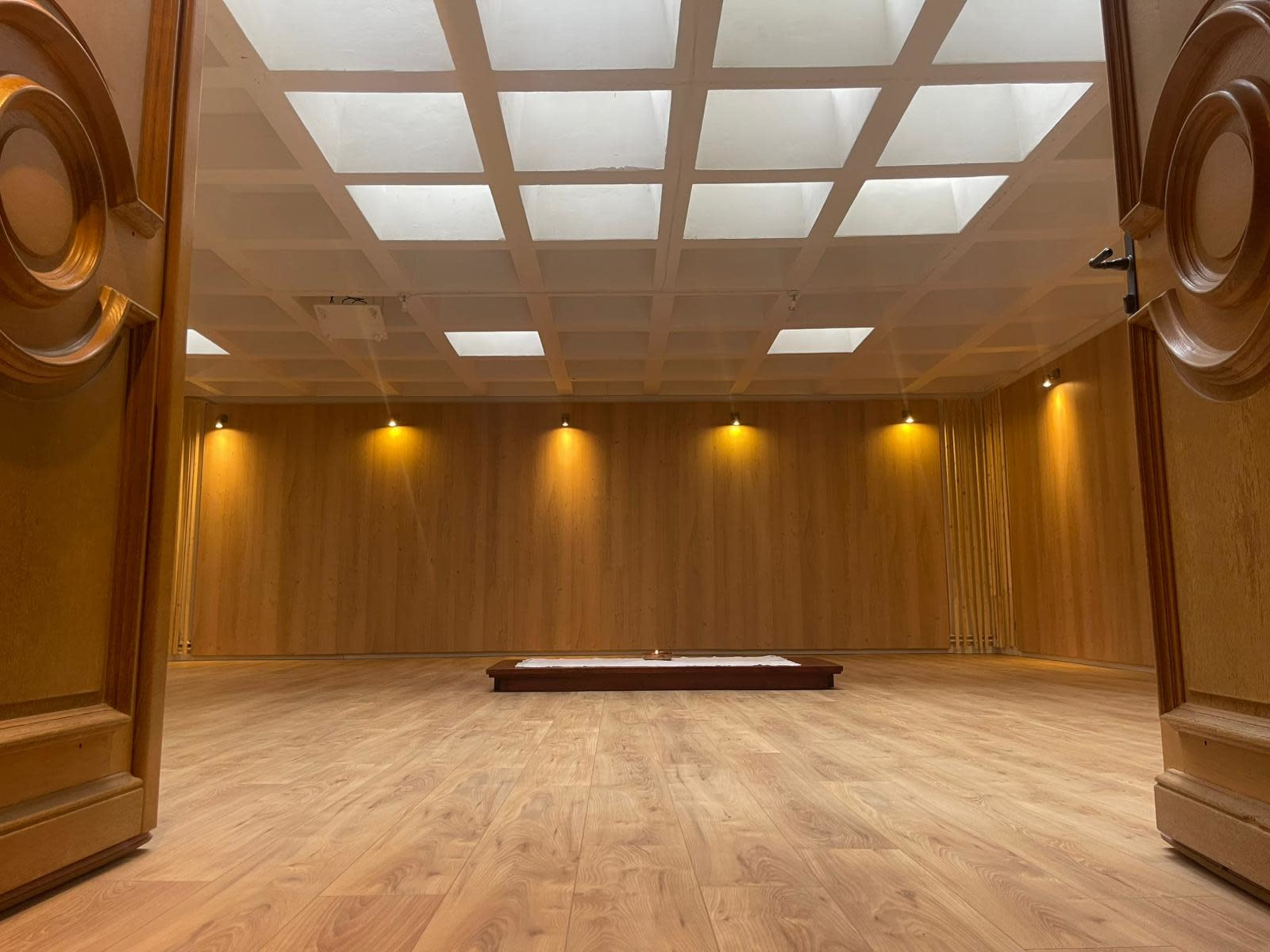 The image shows an empty room with a wooden floor and walls, featuring a coffered ceiling and a small raised platform in the center.