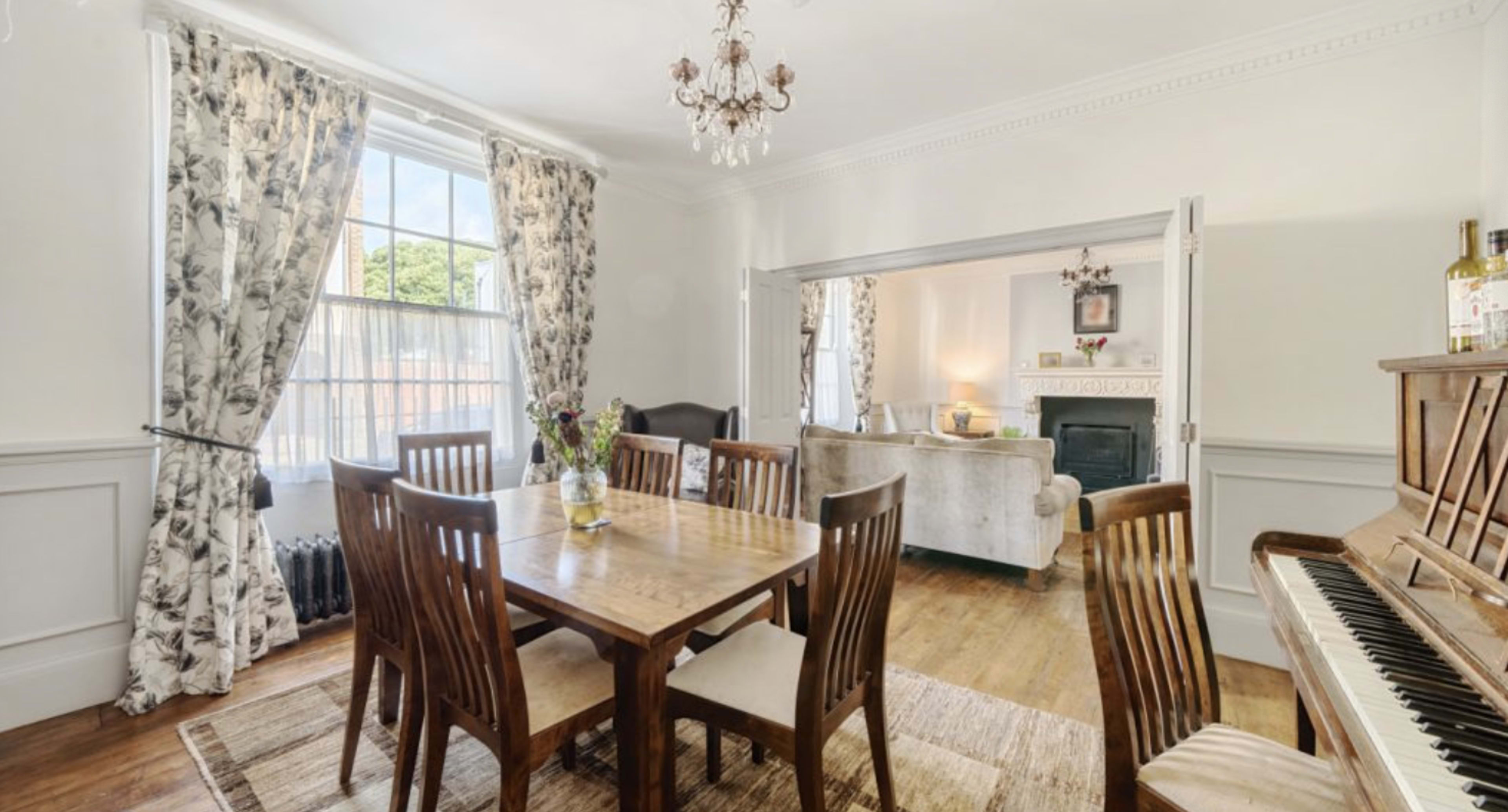 A dining room features a wooden table surrounded by chairs, with large windows draped in floral curtains and a piano in the corner.