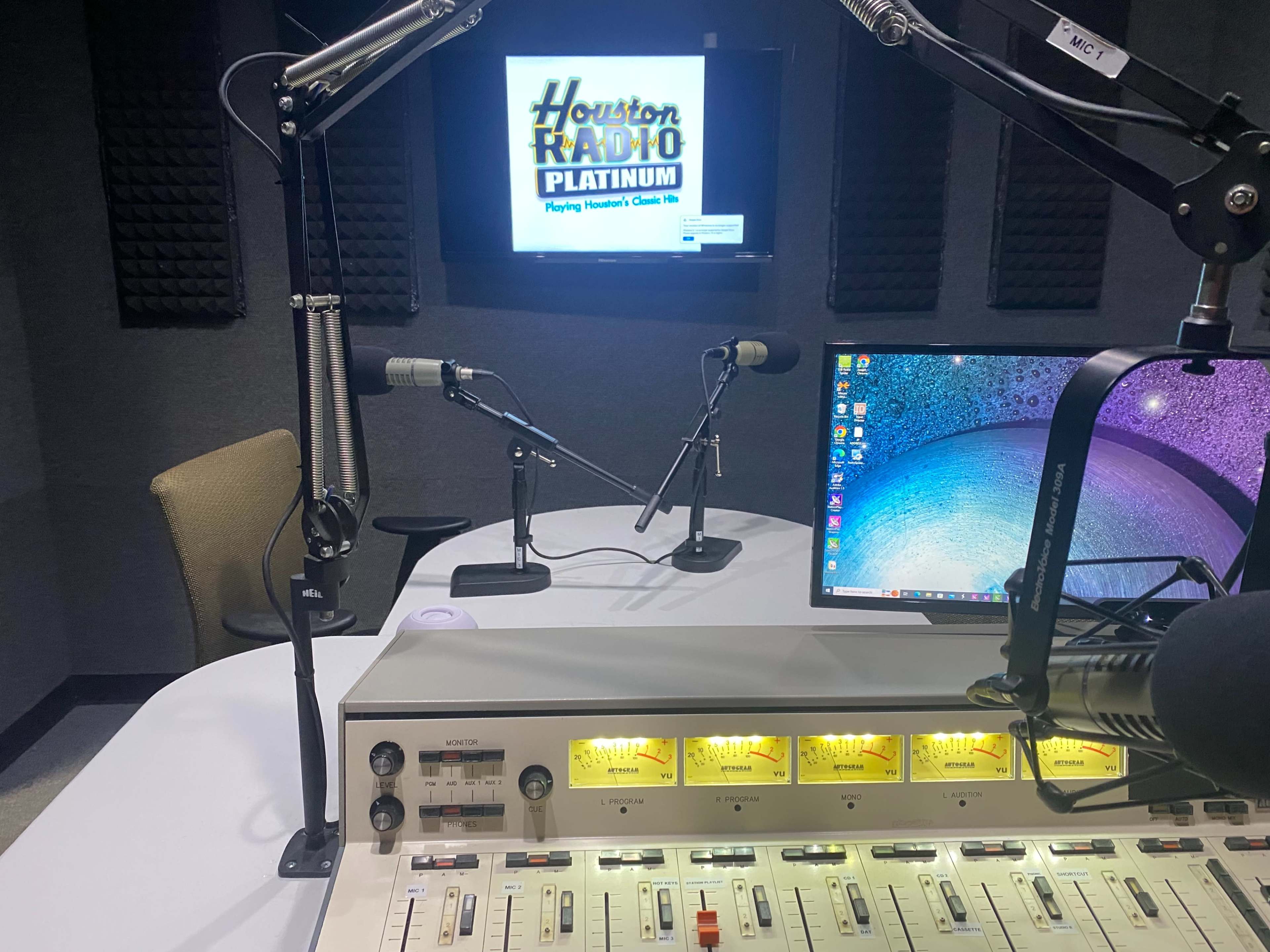 A radio studio with a control panel in the foreground, two microphones, a monitor displaying "Houston Radio Platinum," and a computer.