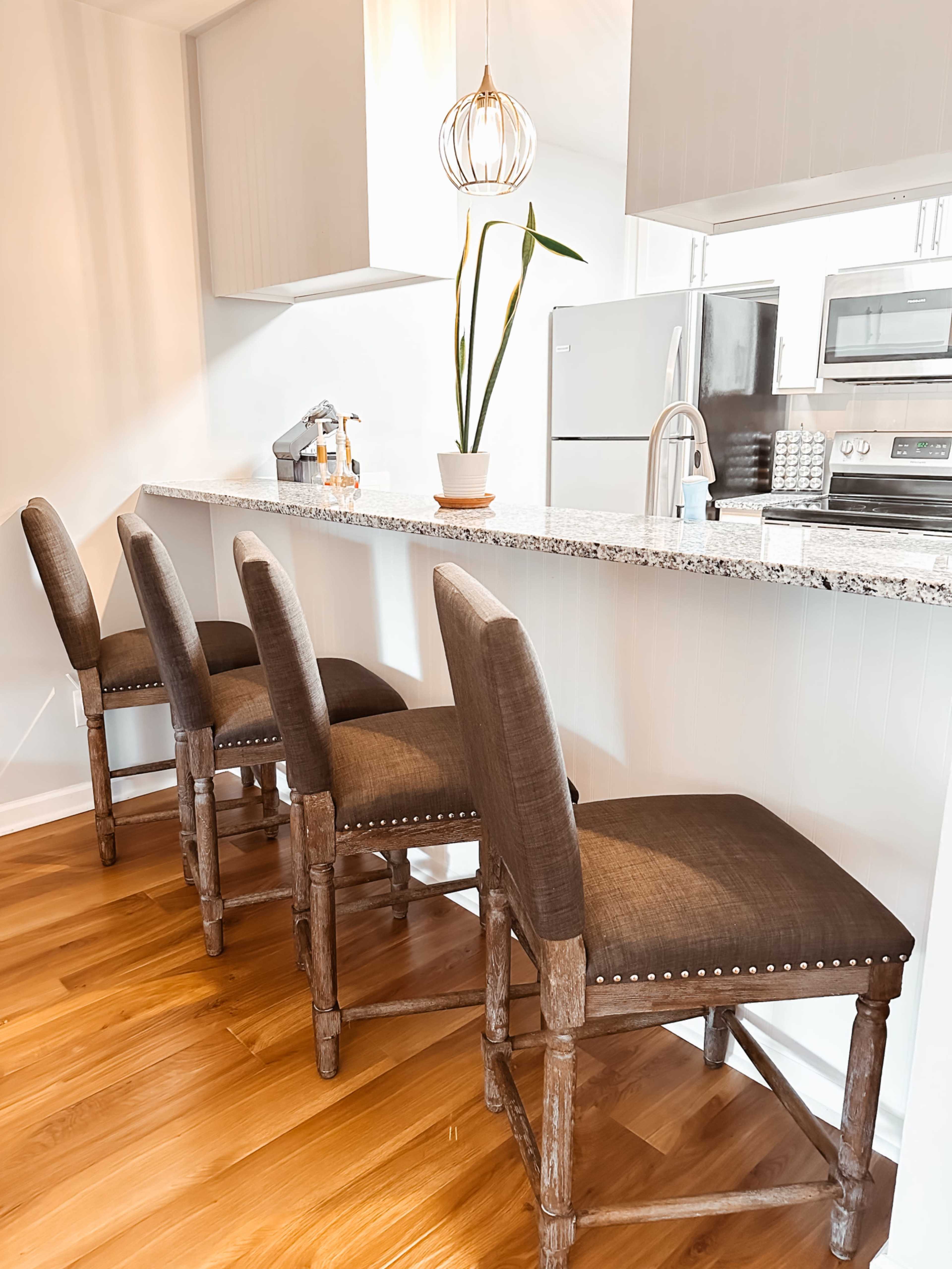 The image shows a modern kitchen bar with four upholstered chairs and a countertop featuring a small potted plant.