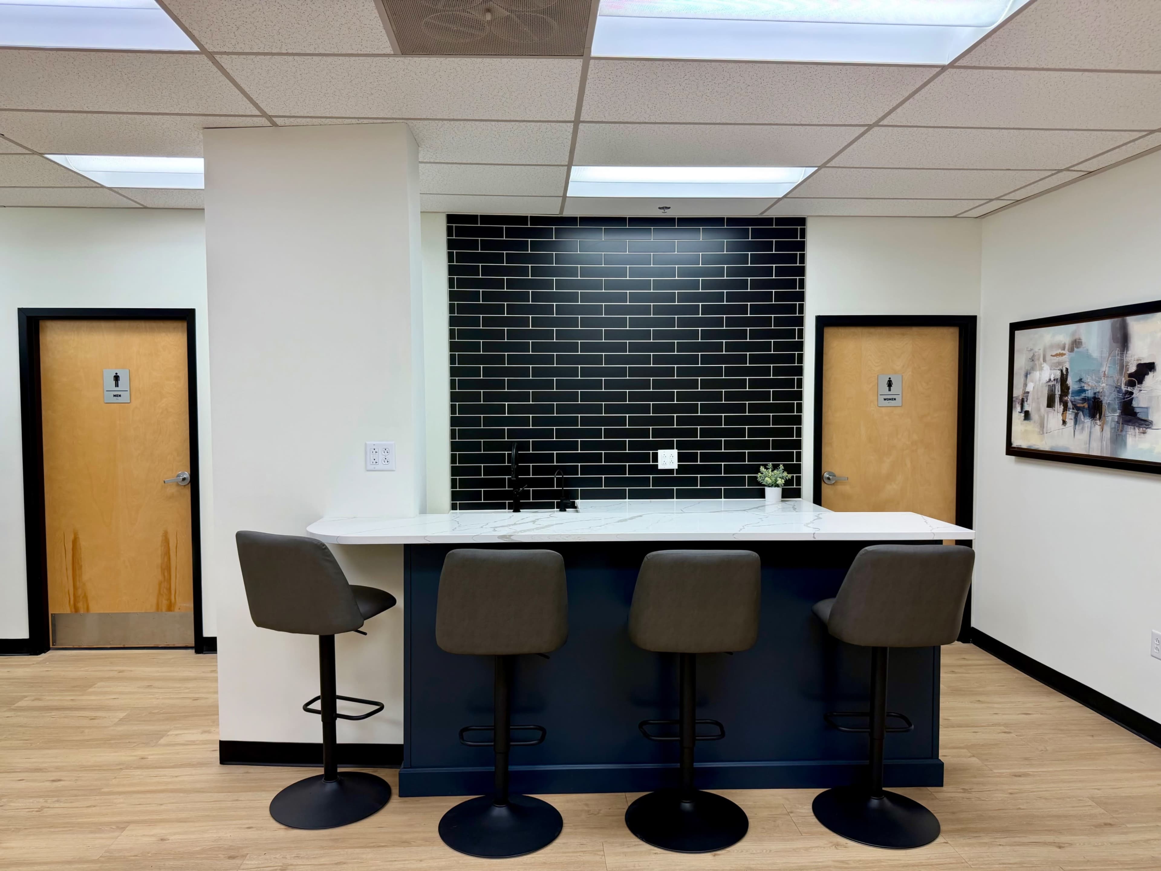 The image shows a modern kitchenette with a bar counter and four high stools, complemented by a dark tiled backsplash and two restroom doors nearby.