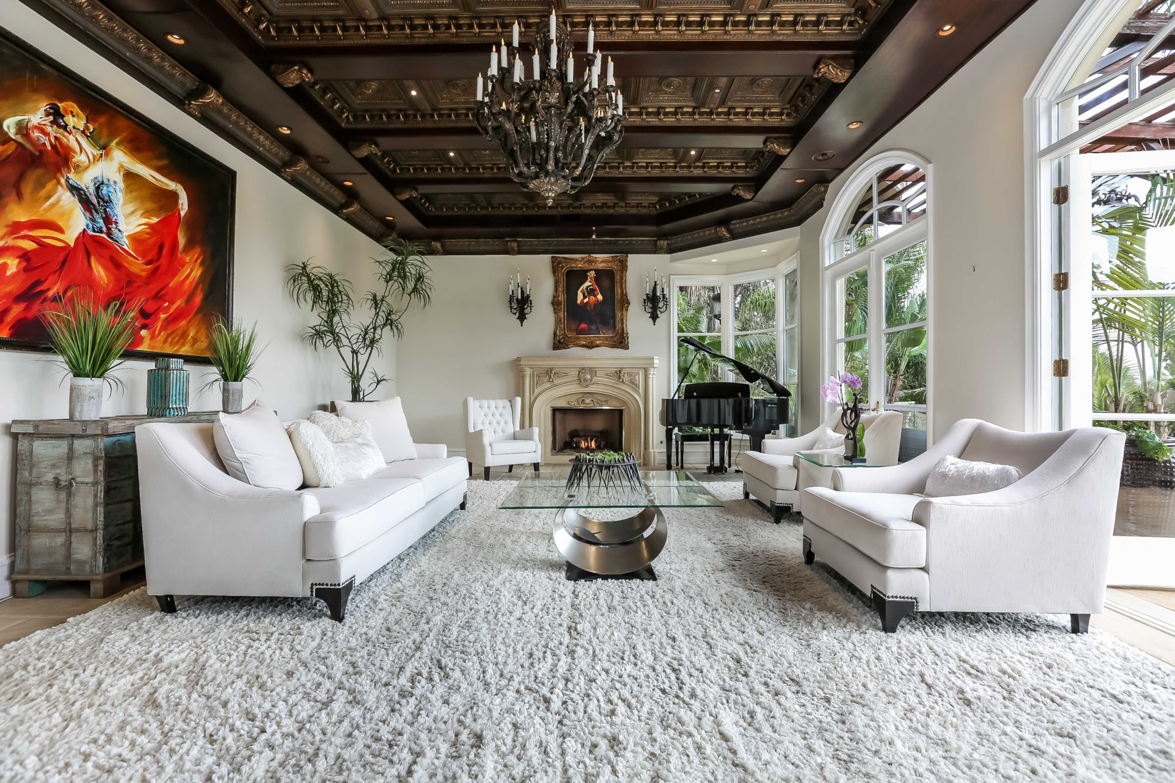 The image shows an elegantly furnished living room with white sofas, a grand piano, and a large chandelier under a decorative ceiling, surrounded by large windows and greenery.