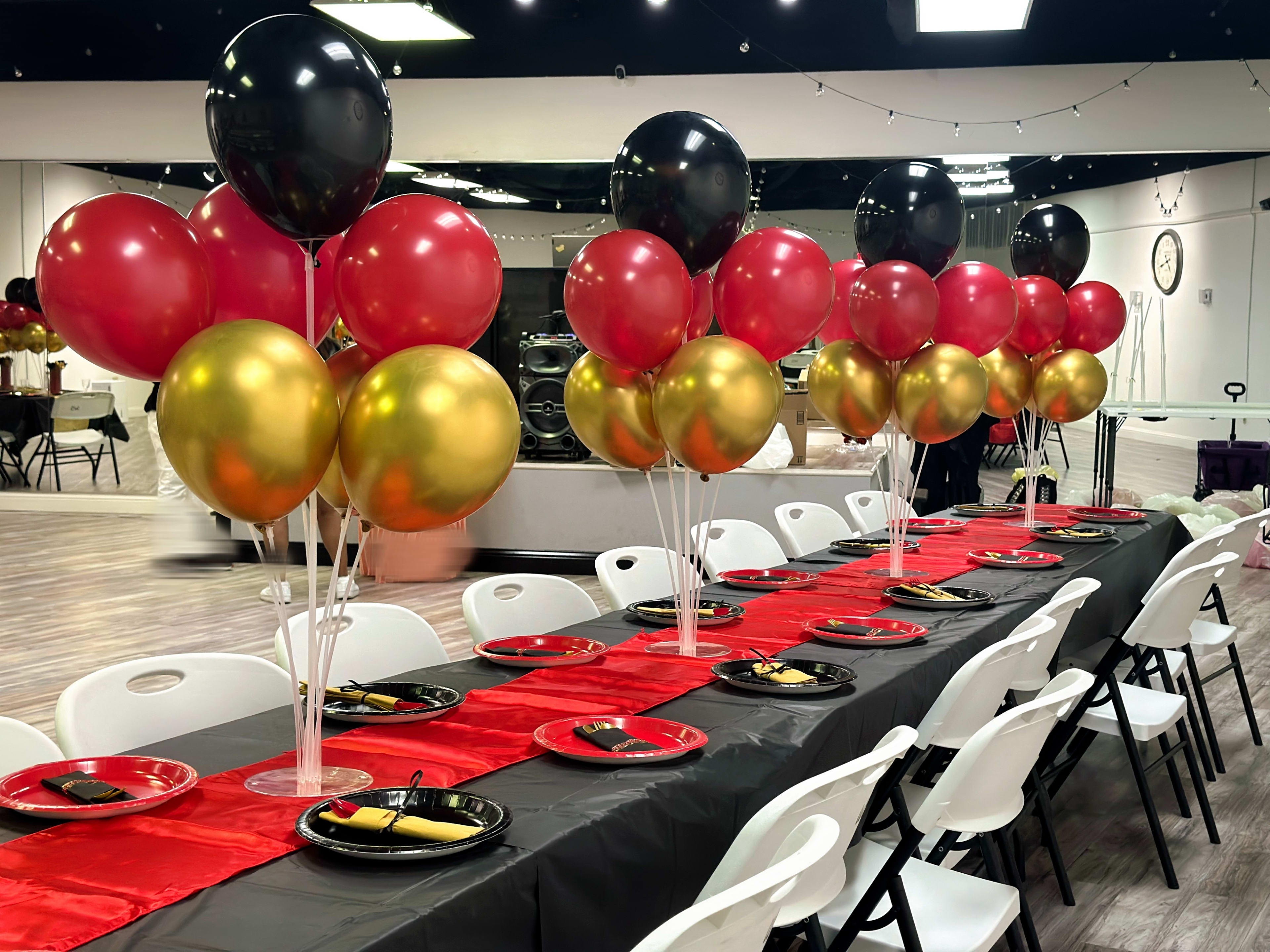 A long banquet table is decorated with red, black, and gold balloons, black tableware, and red table runners in a well-lit event space.