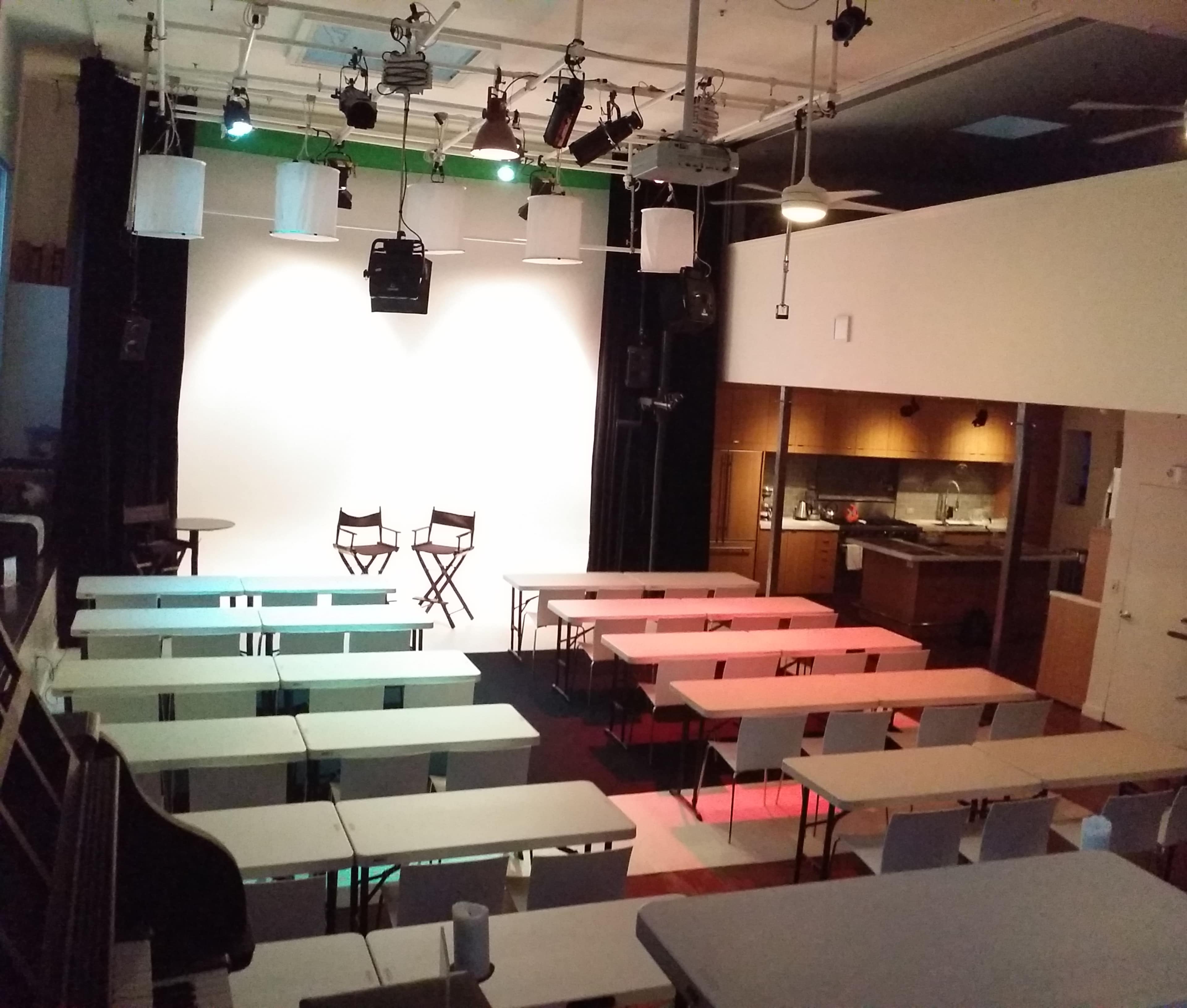 An empty theater or classroom setup with rows of tables and chairs facing a large white screen, flanked by an on-stage area with two director's chairs and a kitchenette in the background.