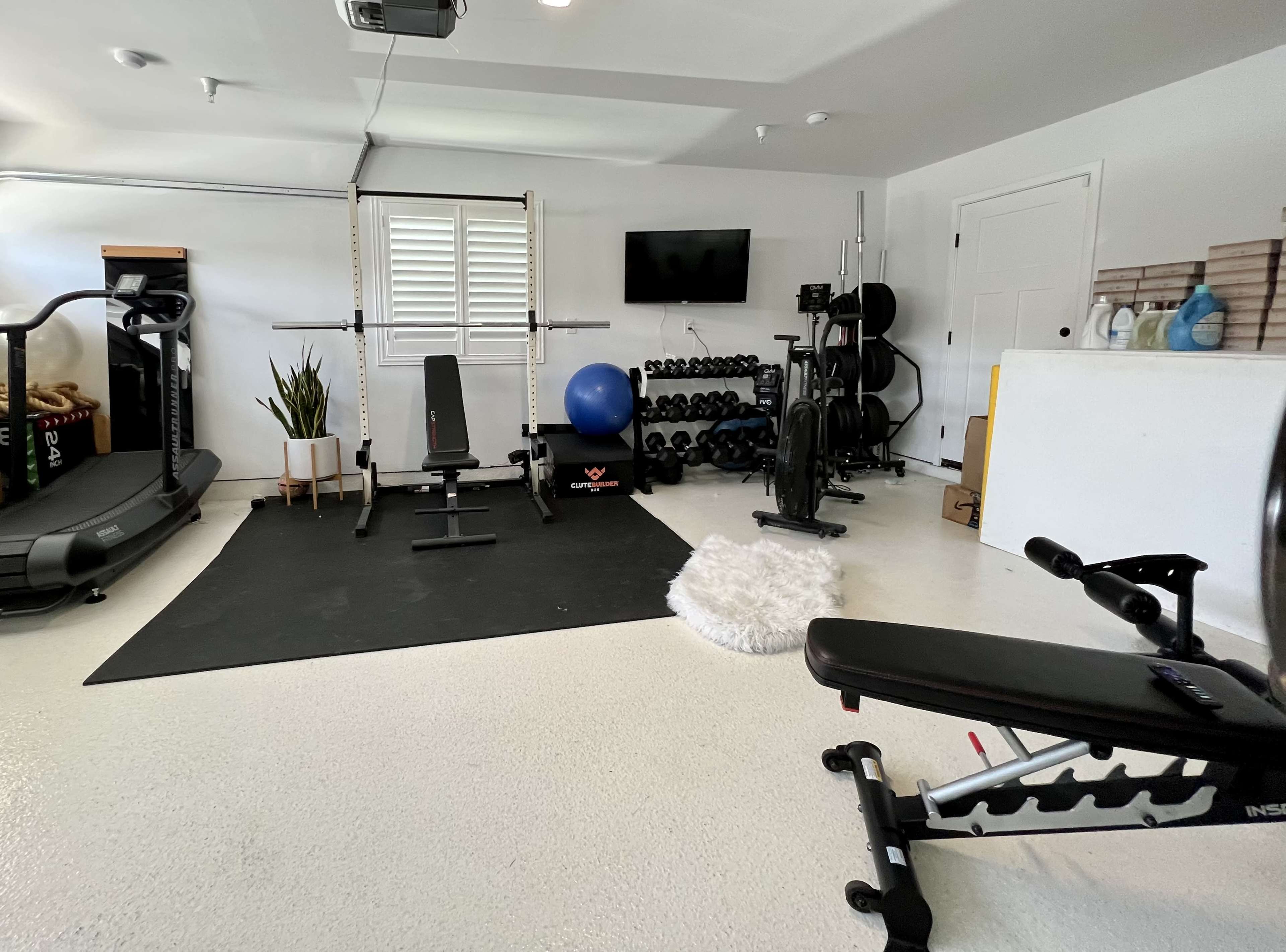 A garage gym setup featuring various exercise equipment, including a treadmill, weight bench, barbell with weights, and a stability ball, all arranged on a rubber mat.
