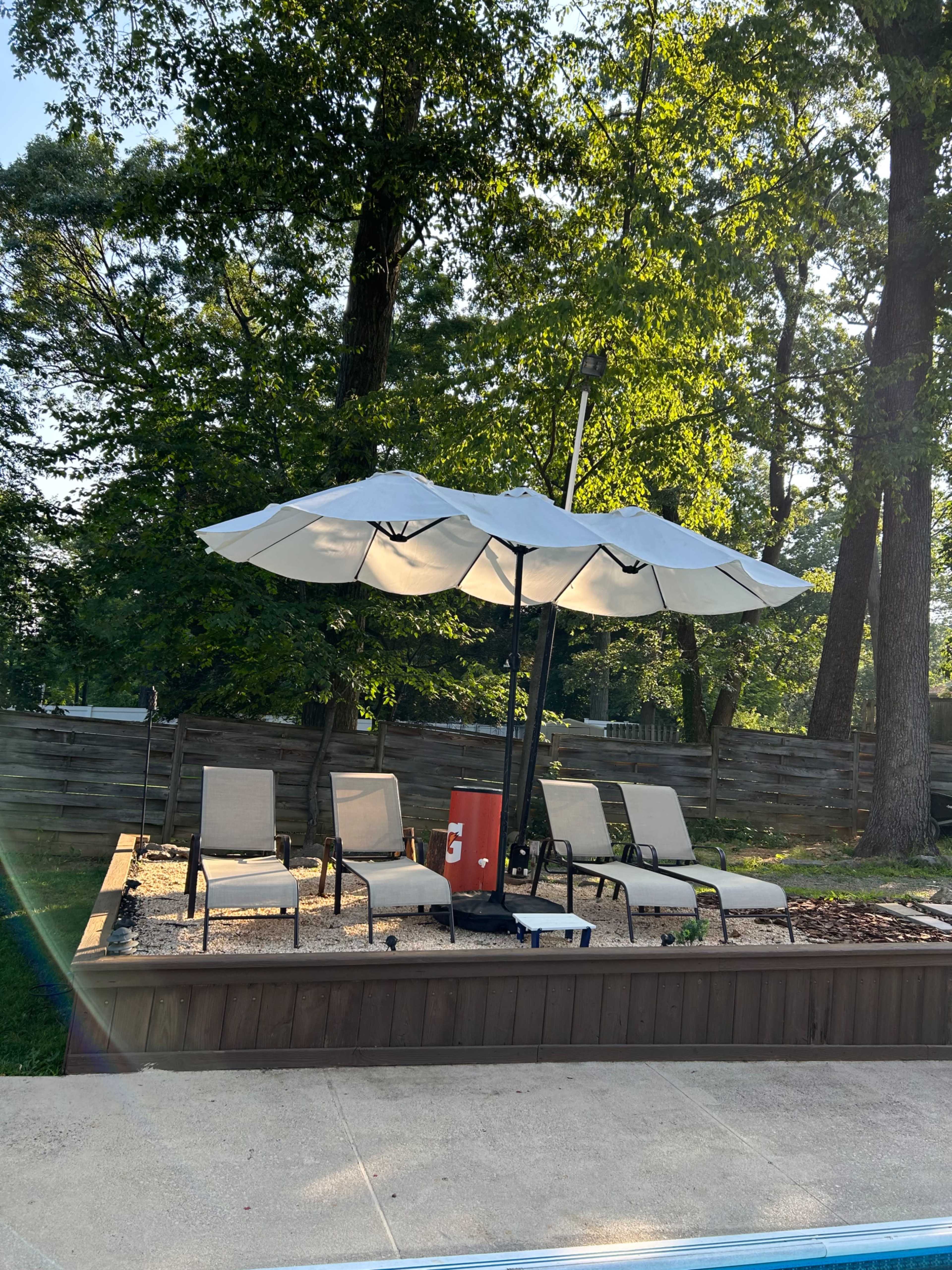 A shaded seating area with lounge chairs and a large umbrella is situated beside a swimming pool, surrounded by trees.