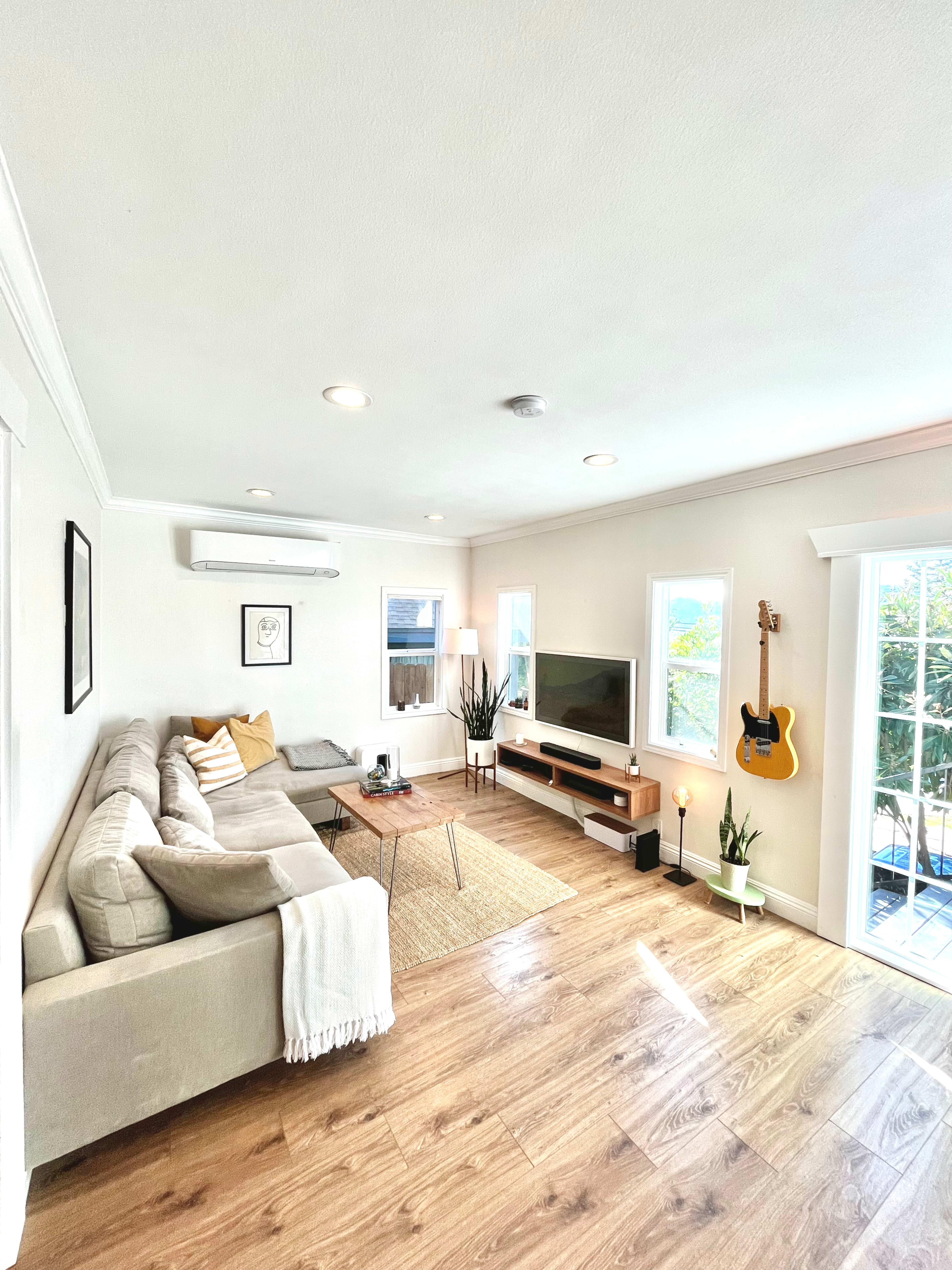 A bright living room features a beige sofa, a wooden coffee table, a television stand with a TV, and large windows overlooking a balcony.