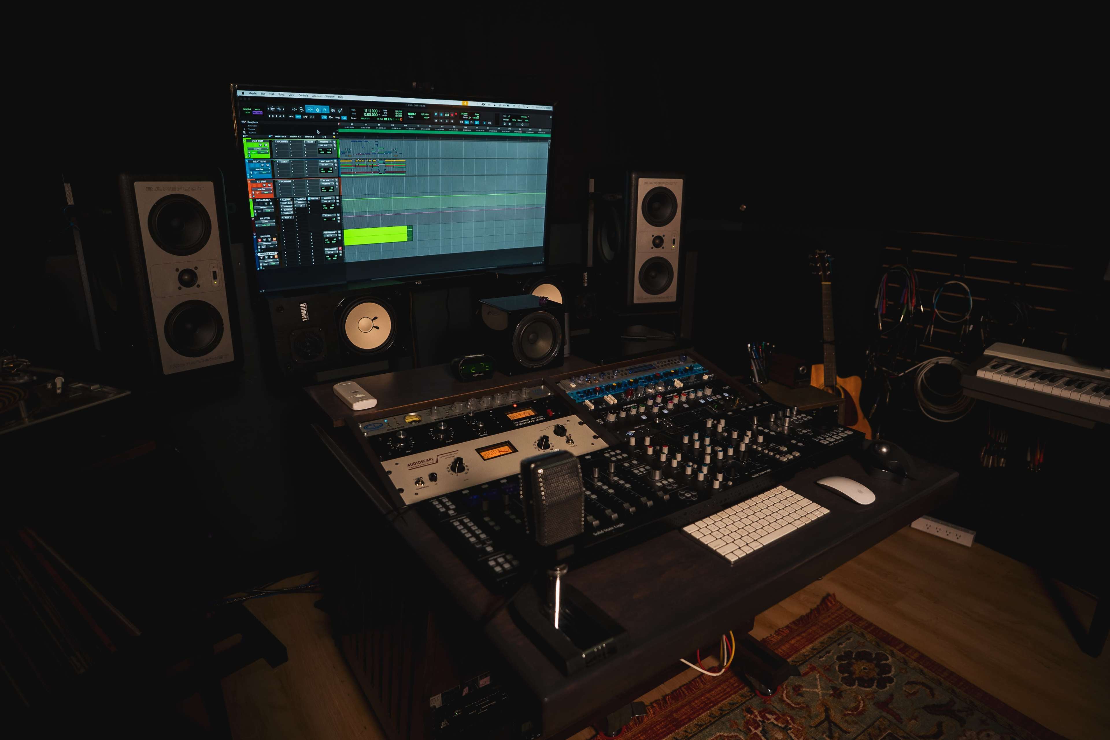 The image shows a music studio with a mixing console, computer monitor displaying audio tracks, and various audio equipment and instruments.